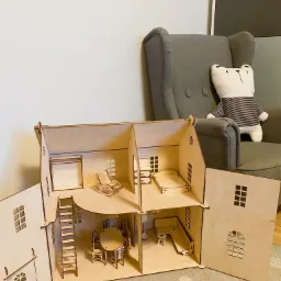 Drewniany domek dla lalek z wyposażeniem, otwarty, z widocznymi meblami w pokojach: krzesło bujane, stół z krzesłami, sofa, łóżko. W tle fotel i pluszowy miś.