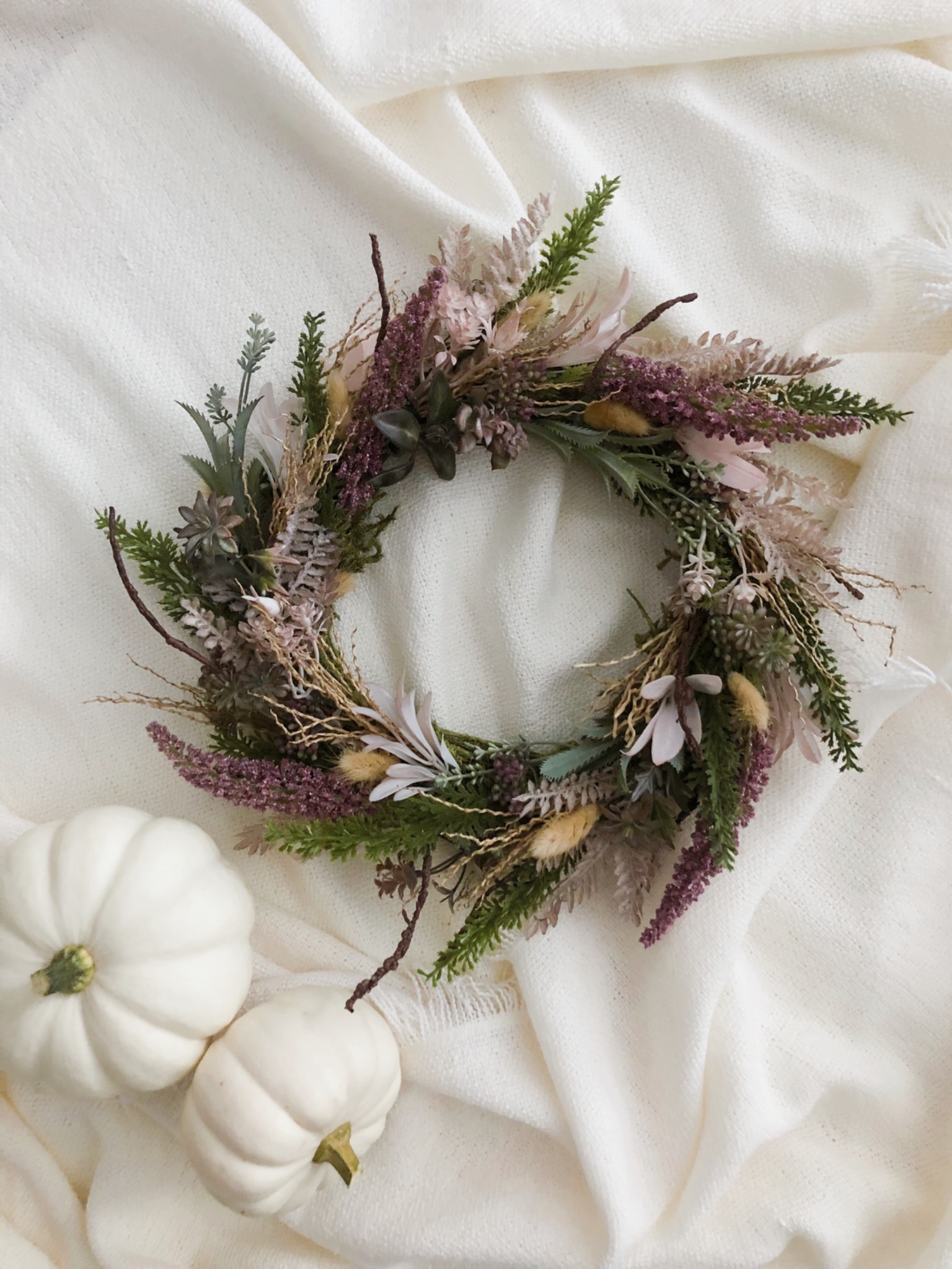 Jesienny wieniec z suszonych kwiatów i gałązek w odcieniach fioletu, zieleni i beżu, ułożony na kremowym, puszystym kocu obok dwóch małych, białych dyń.