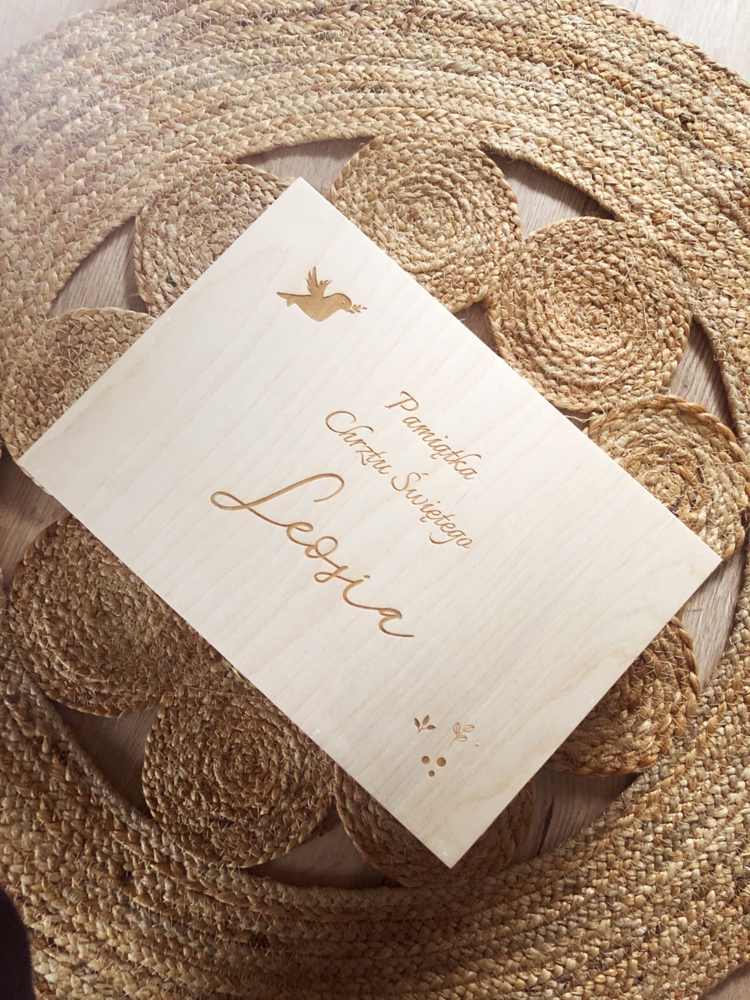 Drewniane pudełko z wygrawerowanym napisem 'Pamiątka Chrztu Świętego Lesia' i motywem gołębia, leżące na okrągłym jutowym dywaniku.