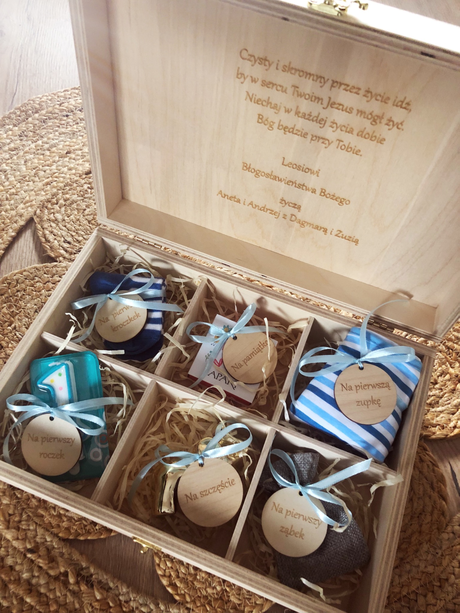 Otwarta drewniana skrzynka z przegródkami wypełnionymi siankiem, zawierająca upominki dla dziecka, takie jak skarpetki, świeczka, materiałowe woreczki i karteczki z napisem 'Na pierwszy roczek'...