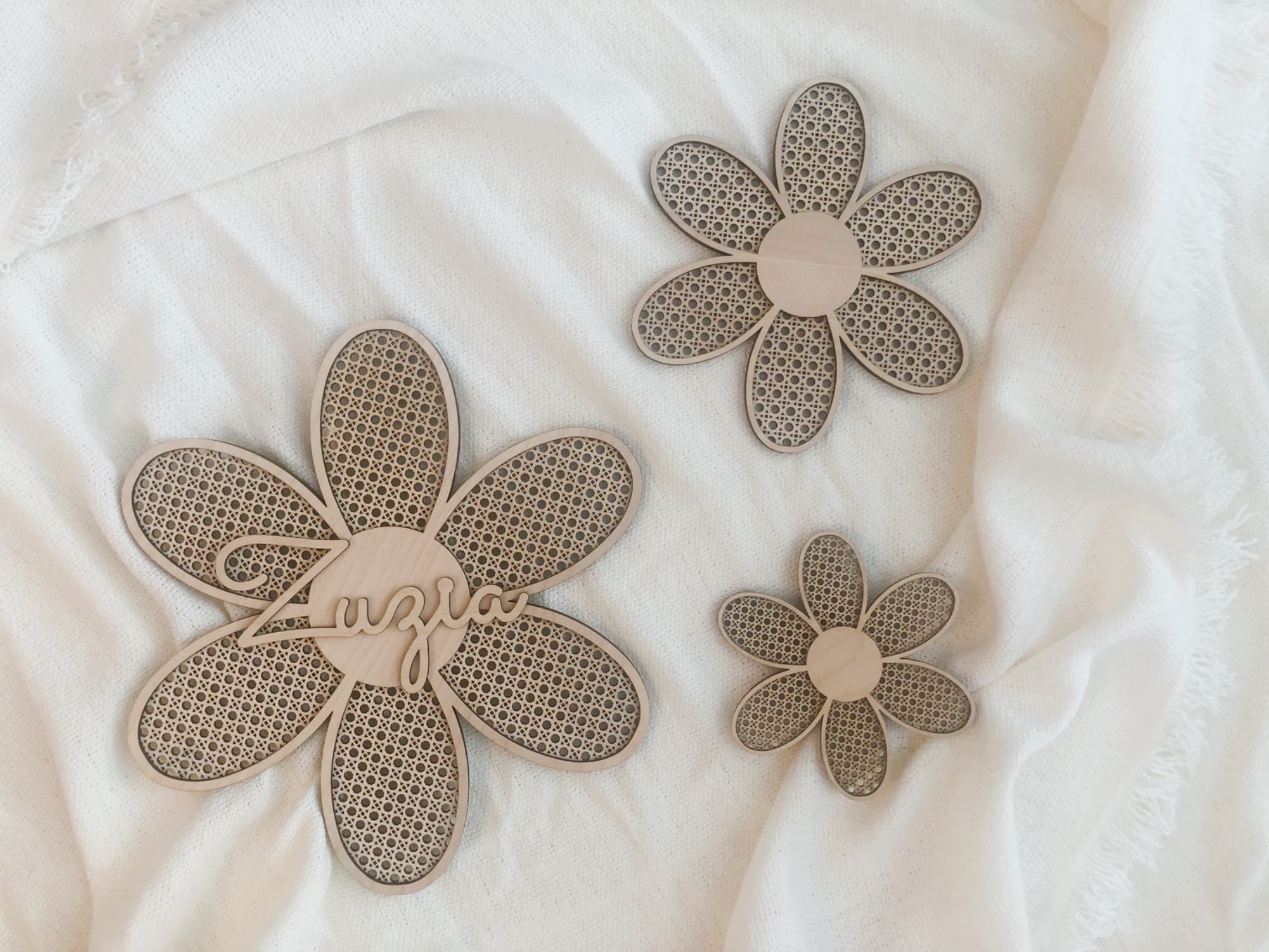 Trzy drewniane ozdoby w kształcie kwiatów z ażurowym wypełnieniem, z czego jeden z napisem 'Zuzia', leżące na białym, puszystym materiale.