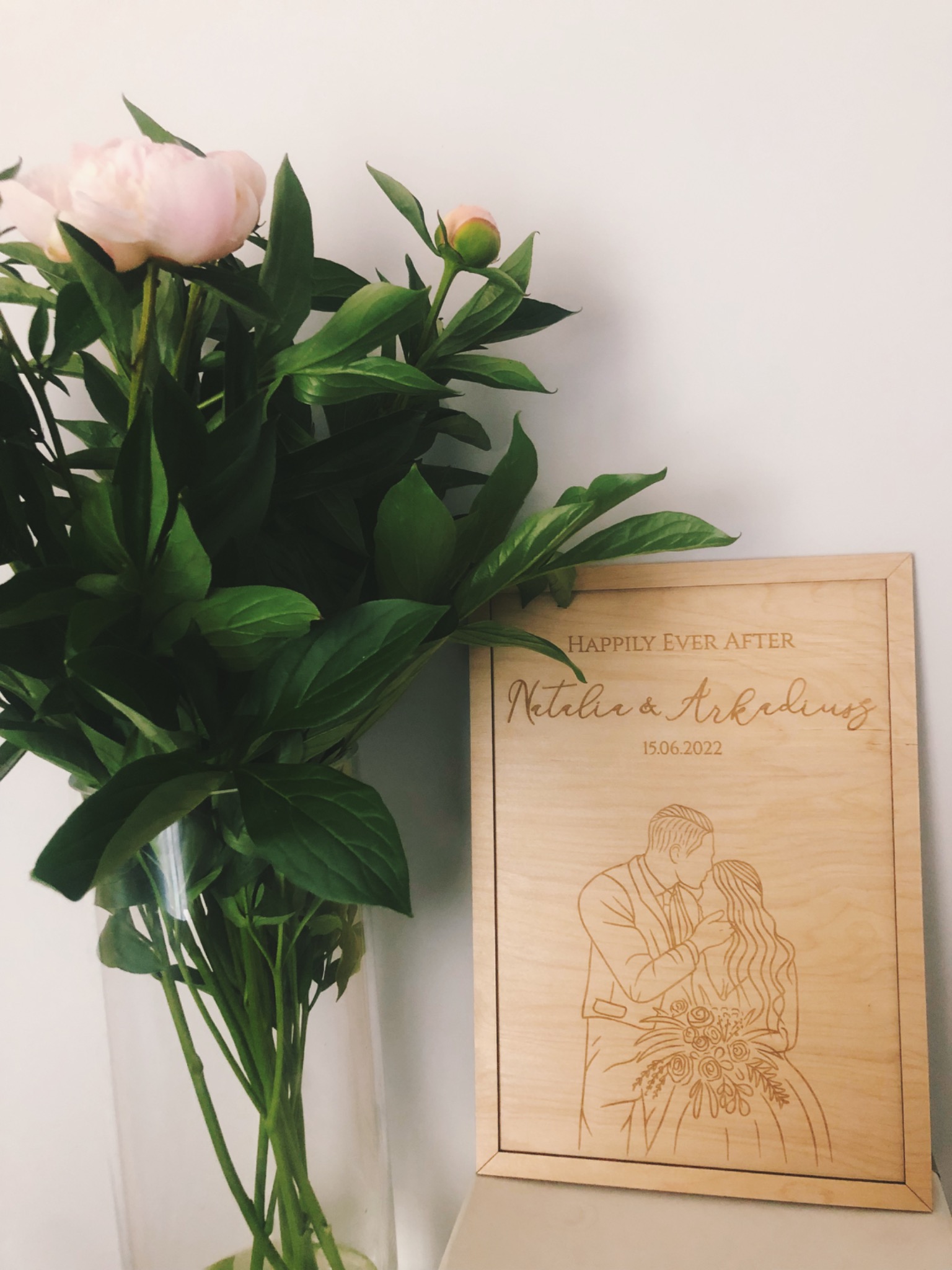 Drewniana tabliczka z wygrawerowanym portretem pary młodej, datą ślubu i napisem 'Happily Ever After', ustawiona obok wazonu z piwoniami.