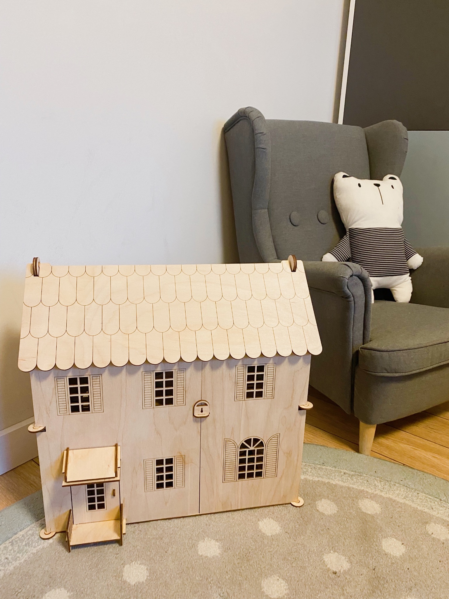 Drewniany domek dla lalek z otwieranymi okiennicami, umieszczony na okrągłym dywanie w kropki, obok szarego fotela z poduszką w kształcie misia.