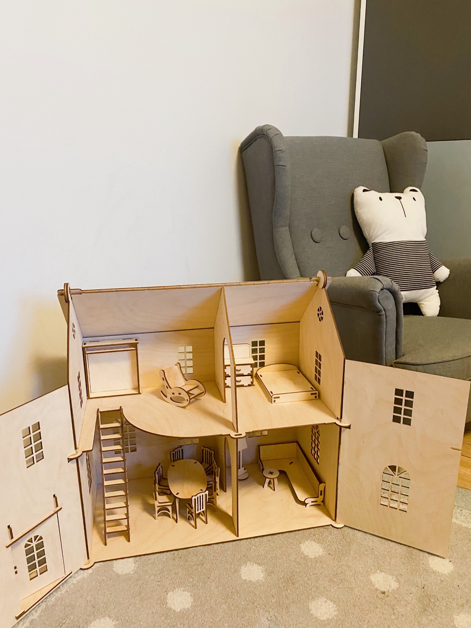 Drewniany domek dla lalek z wyposażeniem, otwarty, z widocznymi meblami w pokojach: krzesło bujane, stół z krzesłami, sofa, łóżko. W tle fotel i pluszowy miś.