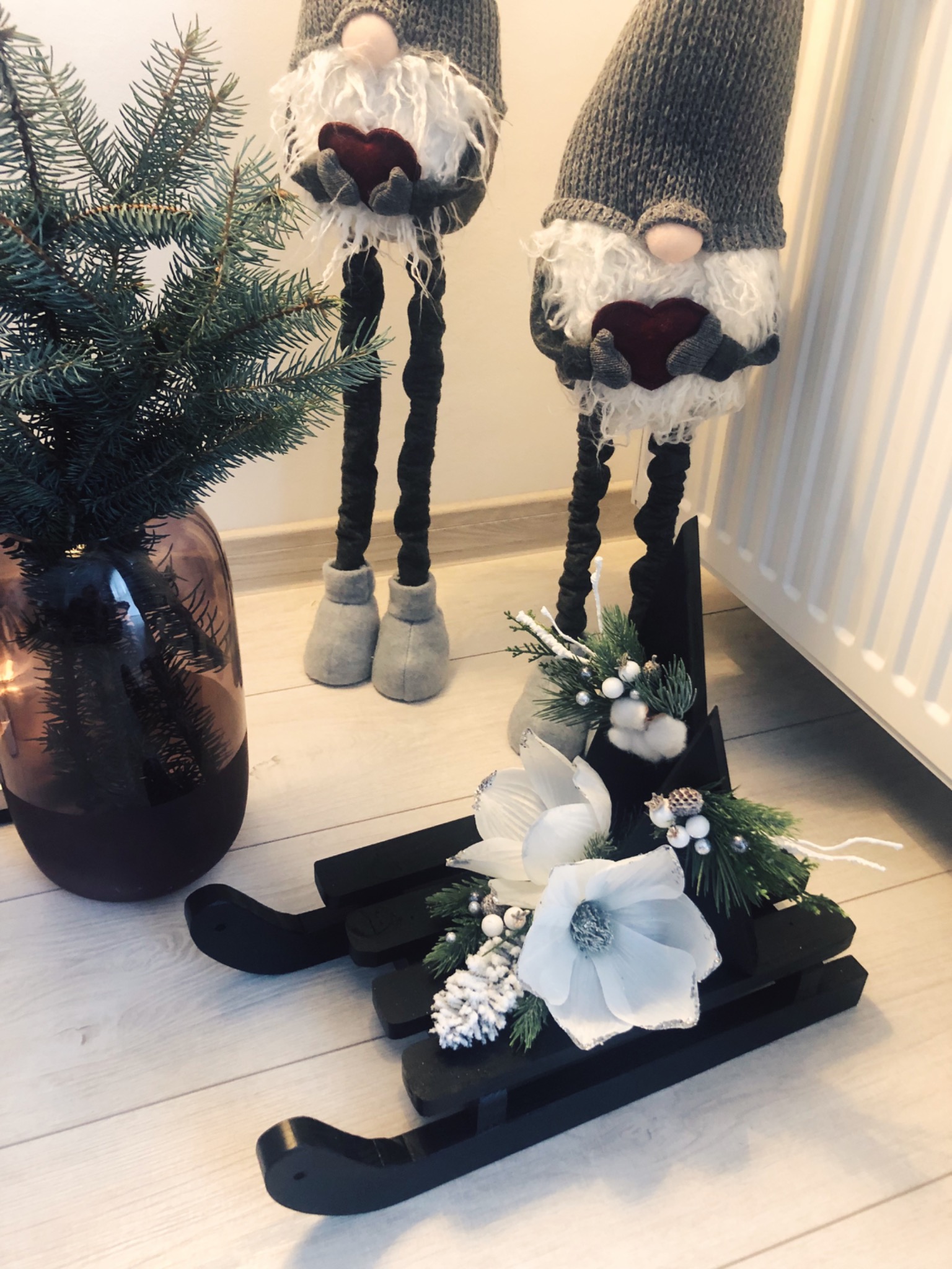 Dekoracja świąteczna: dwa gnomy z szarymi czapkami i białymi brodami trzymające czerwone serca, obok mała choinka w brązowym wazonie i czarne sanie ozdobione białymi kwiatami i gałązkami.