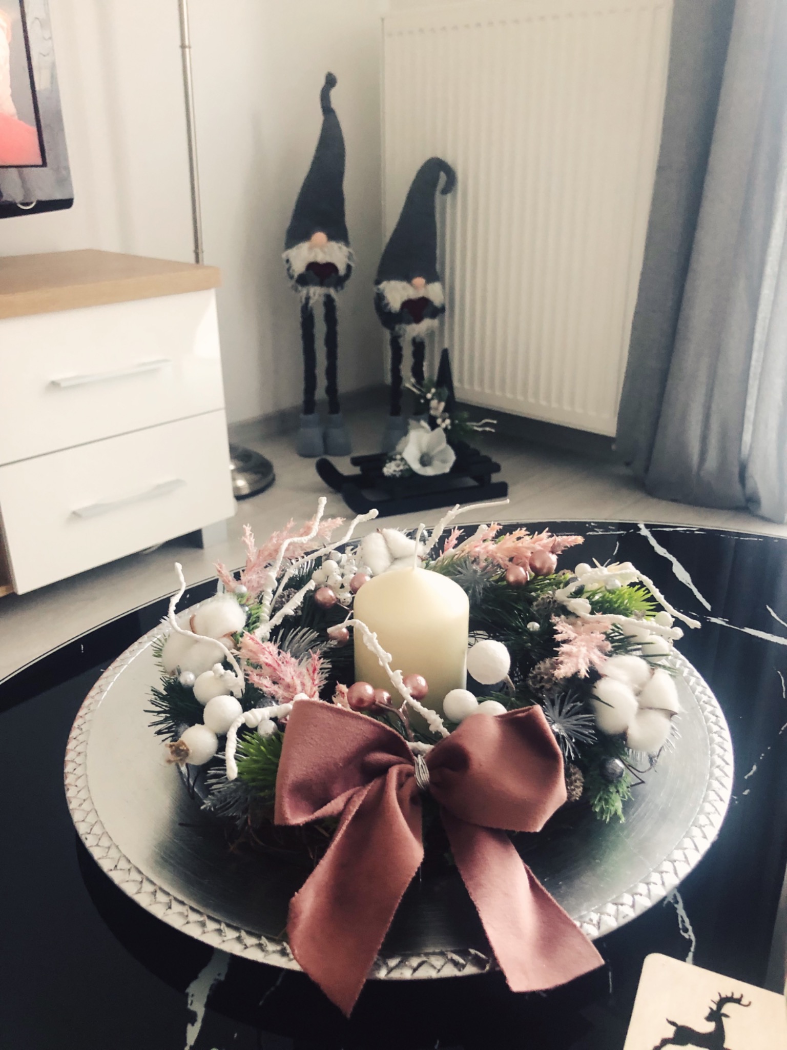 Dekoracyjny wieniec adwentowy ze świecą, ozdobiony różowymi i białymi elementami, leżący na srebrnej tacy na czarnym marmurkowym stole, w tle dwa świąteczne skrzaty.