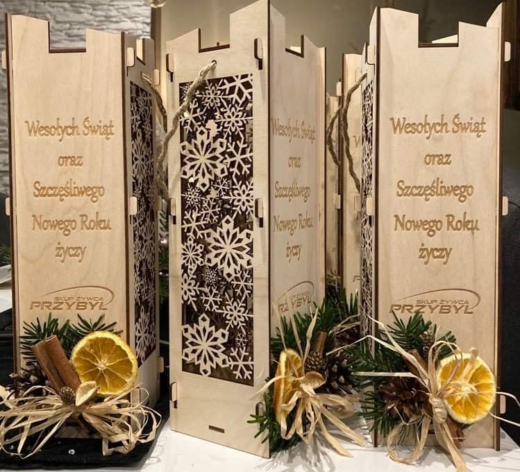 Drewniane skrzynki prezentowe z wyciętymi śnieżynkami i napisem 'Wesołych Świąt oraz Szczęśliwego Nowego Roku', ozdobione suszonymi pomarańczami, cynamonem i szyszkami, z widocznym logo 'Skup Żywca...