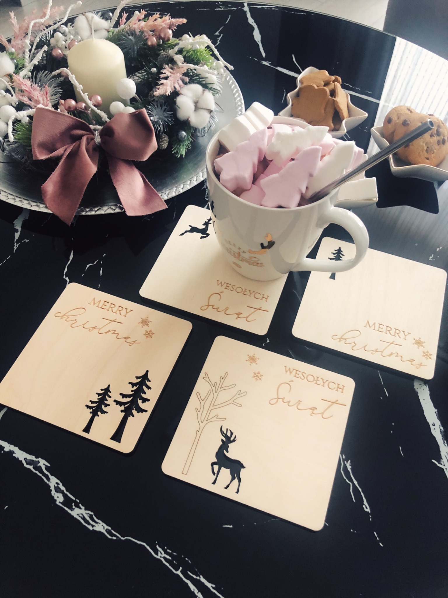 Świąteczny stroik z kremową świecą i kokardą, kubek z piankami, pierniki i podkładki z napisem 'Wesołych Świąt' i motywami świątecznymi na czarnym marmurowym stole.