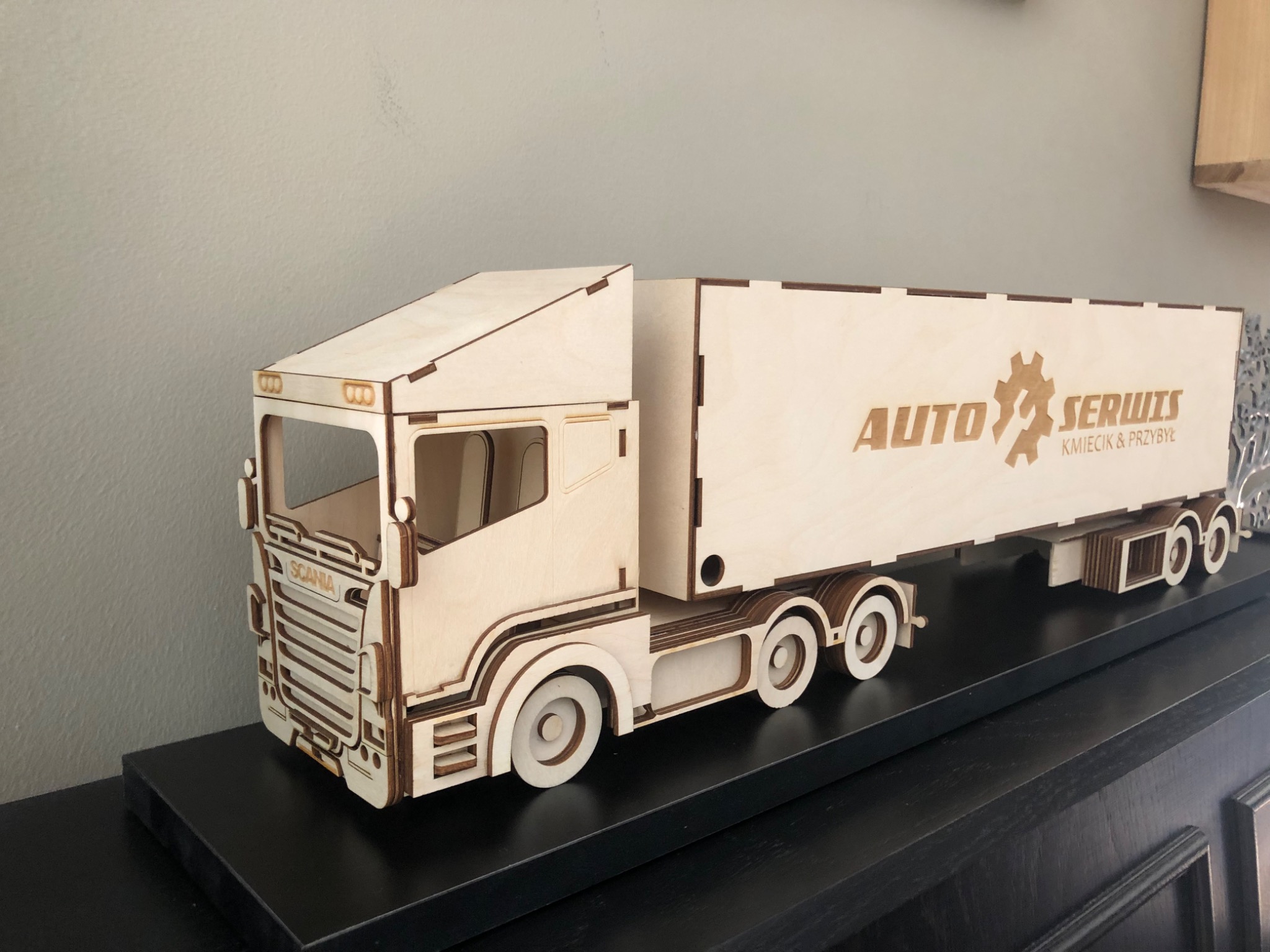 Drewniany model ciężarówki SCANIA z logo 'Auto Serwis Kmiecik & Przybył' na naczepie, stojący na czarnym regale.