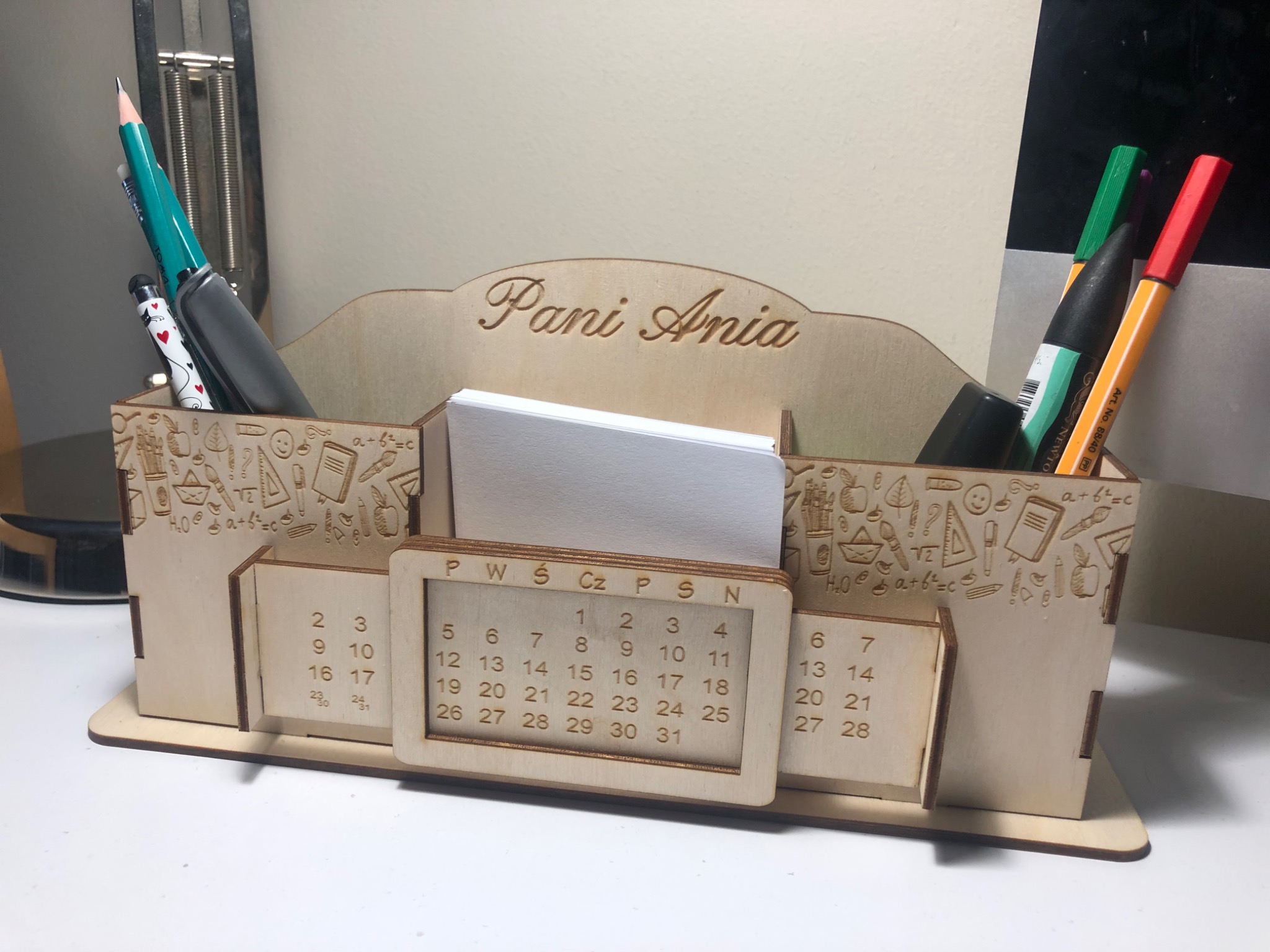 Drewniany organizer biurkowy z kalendarzem, grawerowanymi wzorami szkolnymi i napisem 'Pani Ania', wypełniony długopisami, ołówkami i karteczkami.