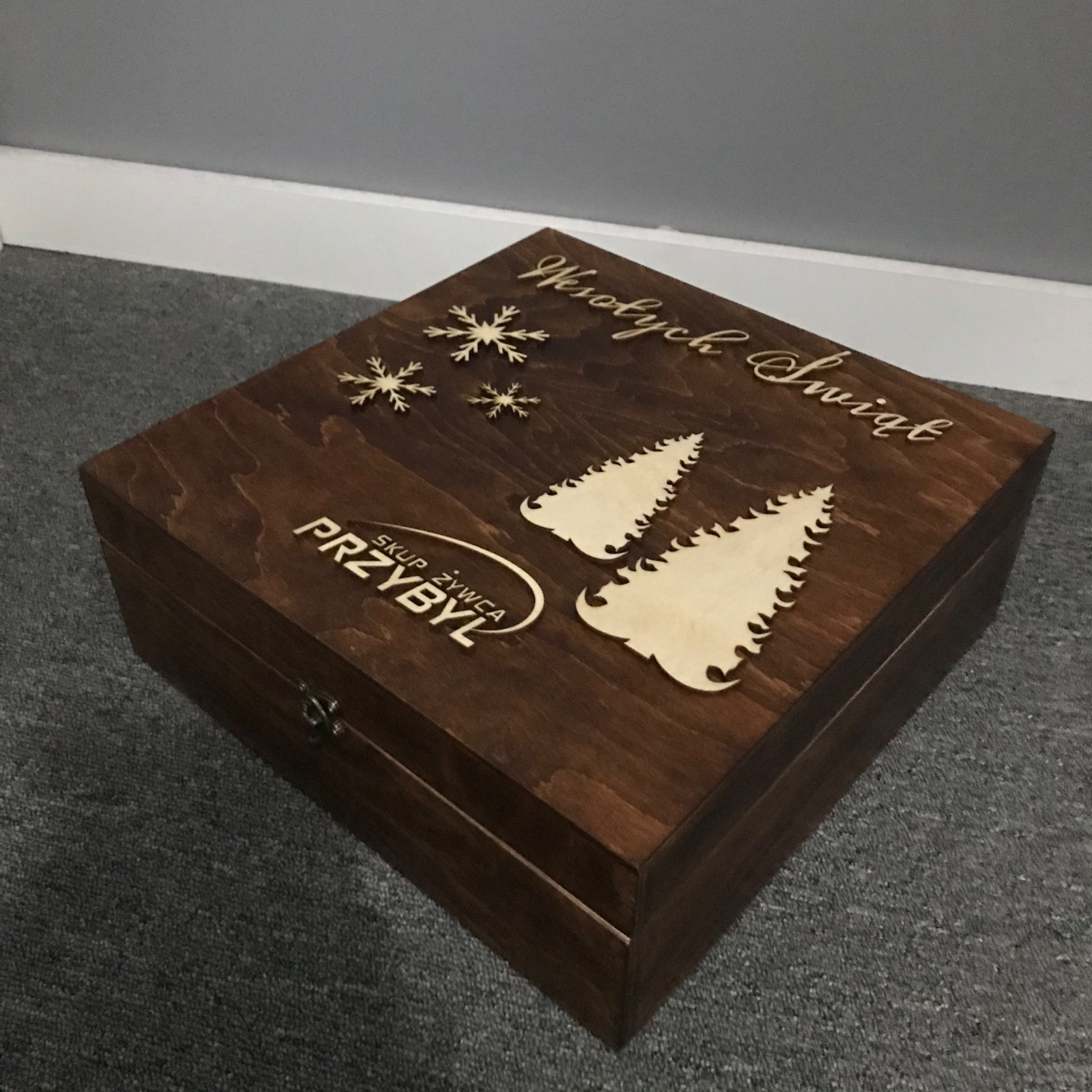 Ciemnobrązowe, drewniane pudełko prezentowe z wygrawerowanymi laserowo śnieżynkami, drzewkami choinkowymi i napisem 'Wesołych Świąt' oraz logo firmy 'Skup Żywca Przybyl'.