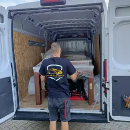 Pracownik firmy transportowej w czarnej koszulce z logo 'Stodulski Trans' ładuje stół owinięty folią do wnętrza białego busa z drewnianą zabudową.