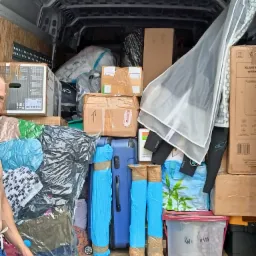 Wnętrze furgonetki załadowane kartonami, workami próżniowymi z ubraniami, niebieską walizką i plastikowym pojemnikiem z napisem 'KWI KIA', częściowo widoczna ręka osoby po lewej stronie.