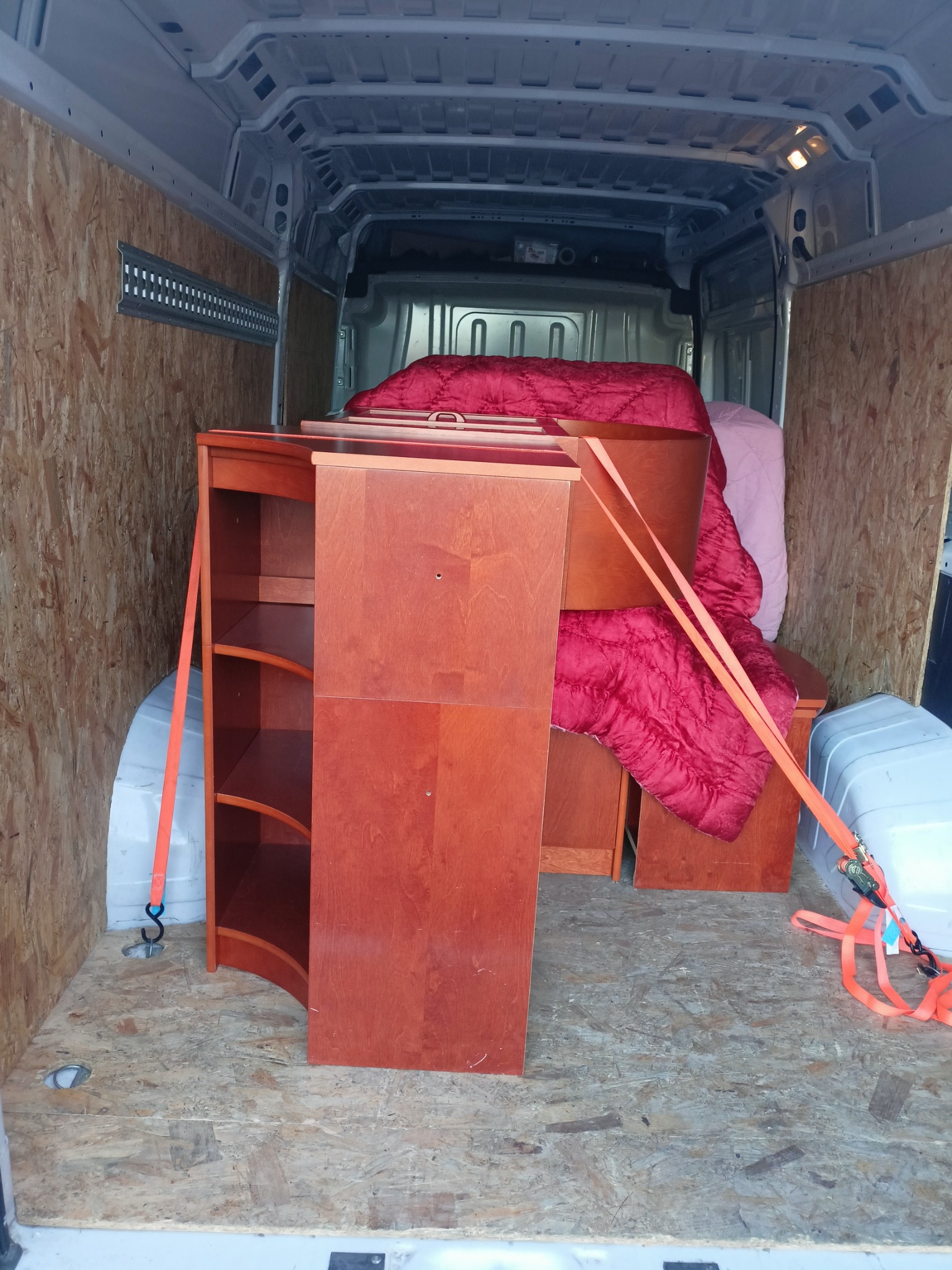 Wnętrze furgonetki z zabezpieczonymi meblami: regał z półkami i inne elementy przykryte czerwoną kołdrą, spięte pomarańczowymi pasami transportowymi.