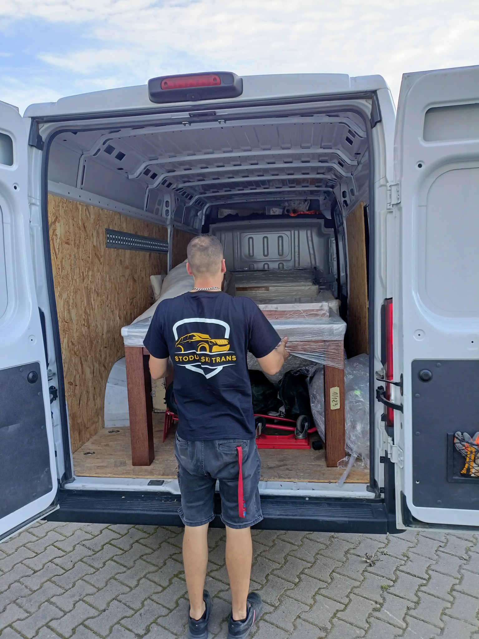 Pracownik firmy transportowej w czarnej koszulce z logo 'Stodulski Trans' ładuje stół owinięty folią do wnętrza białego busa z drewnianą zabudową.