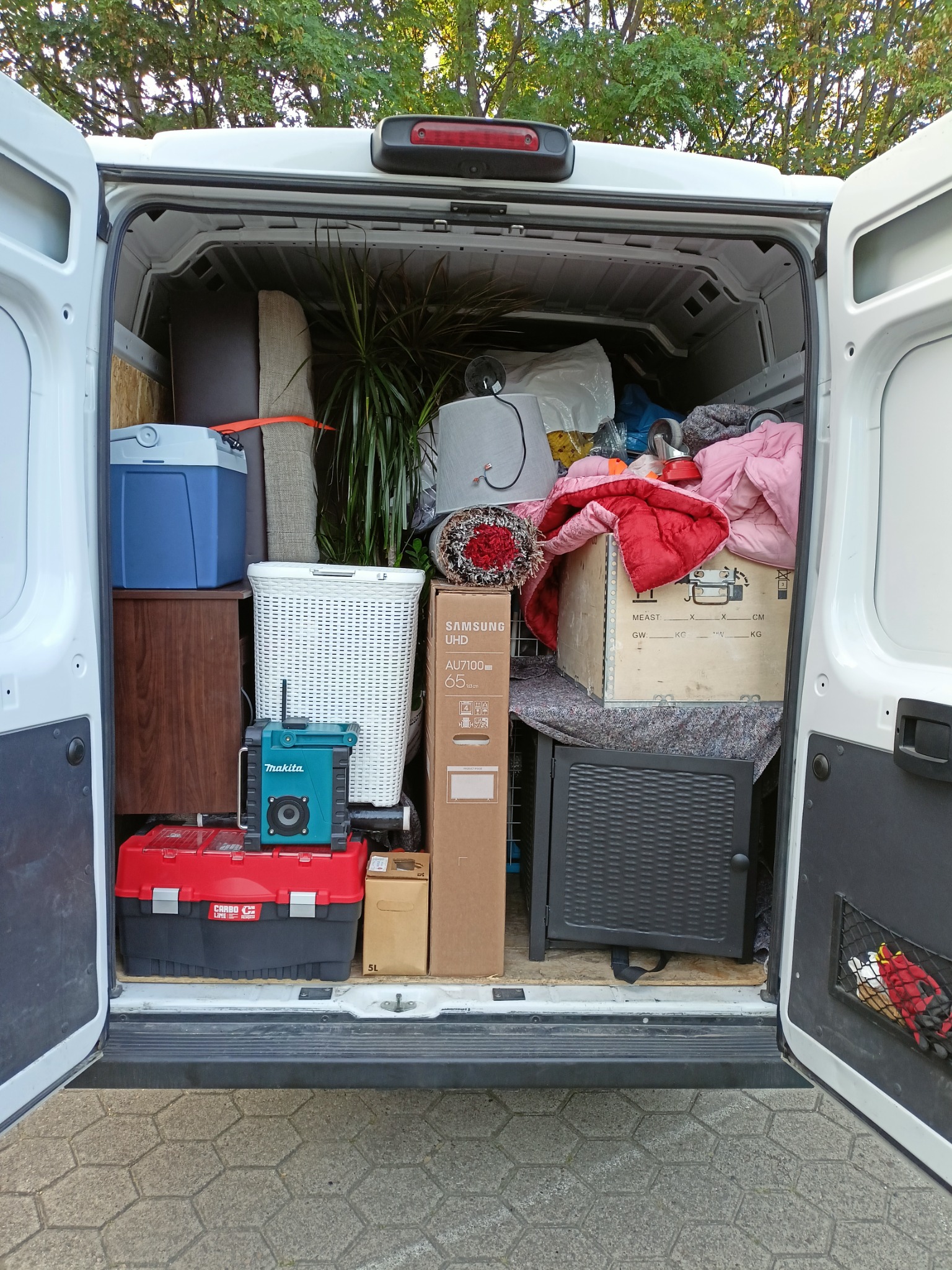 Wnętrze białego busa załadowane różnorodnymi przedmiotami podczas przeprowadzki: meble, kartony, roślina doniczkowa, narzędzia Makita i skrzynka narzędziowa Carbo Line, widoczny karton...