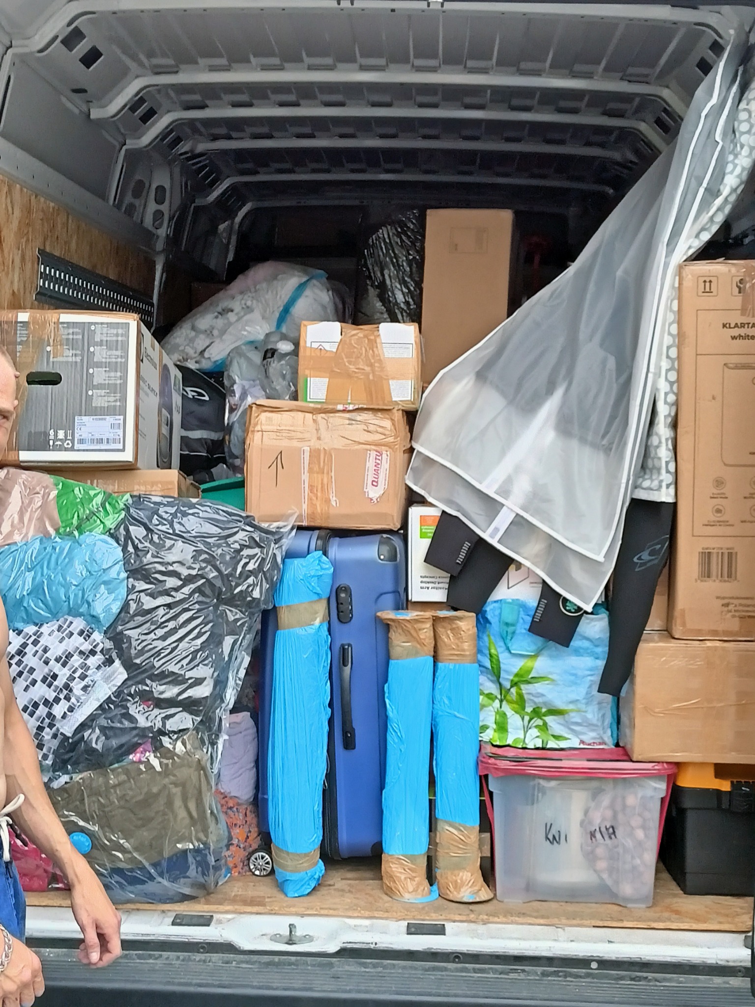 Wnętrze furgonetki załadowane kartonami, workami próżniowymi z ubraniami, niebieską walizką i plastikowym pojemnikiem z napisem 'KWI KIA', częściowo widoczna ręka osoby po lewej stronie.