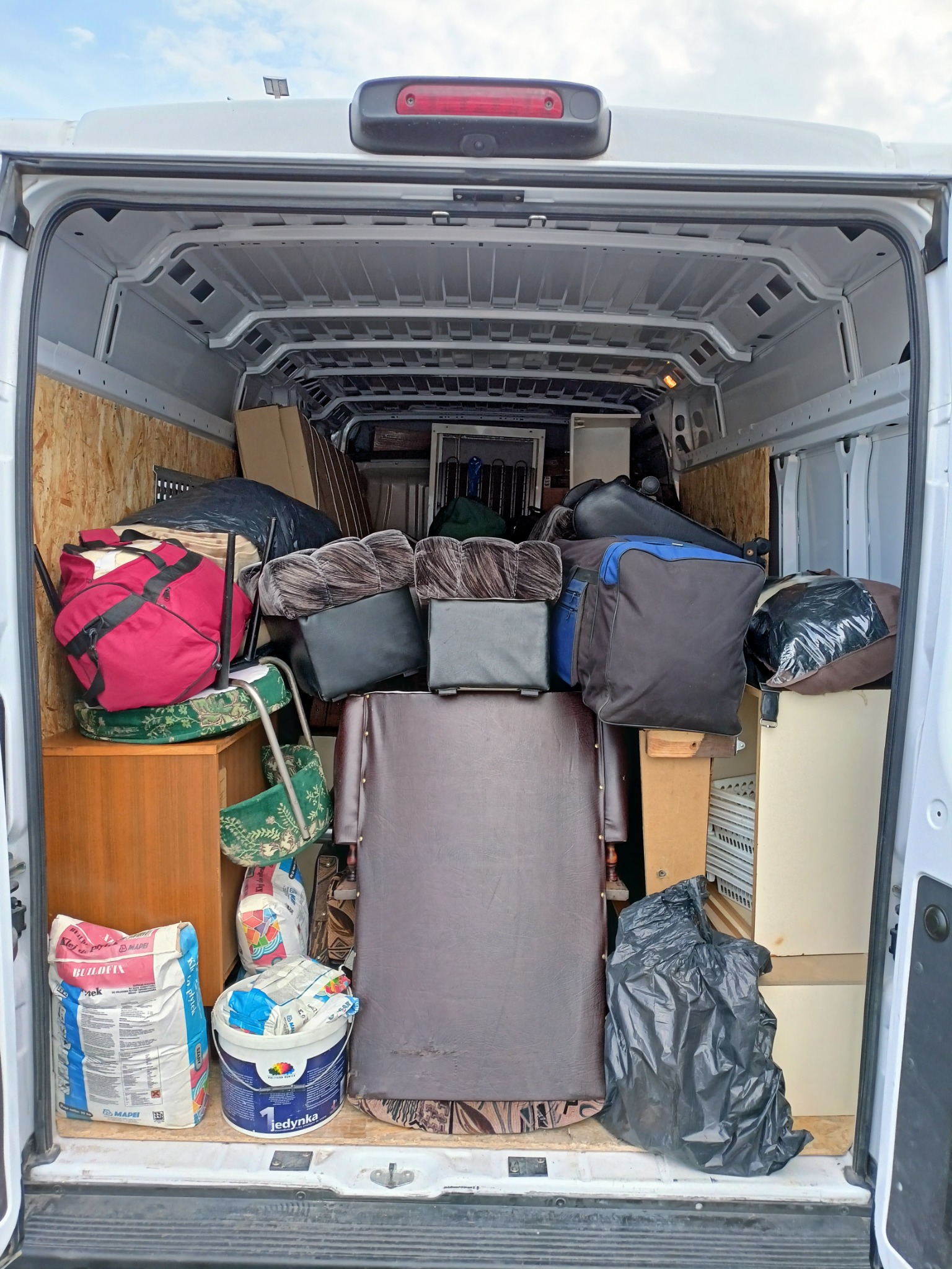 Wnętrze białego busa załadowane meblami, torbami i materiałami budowlanymi, w tym workiem kleju 'Budfix' i wiadrem farby 'Jedynka'.