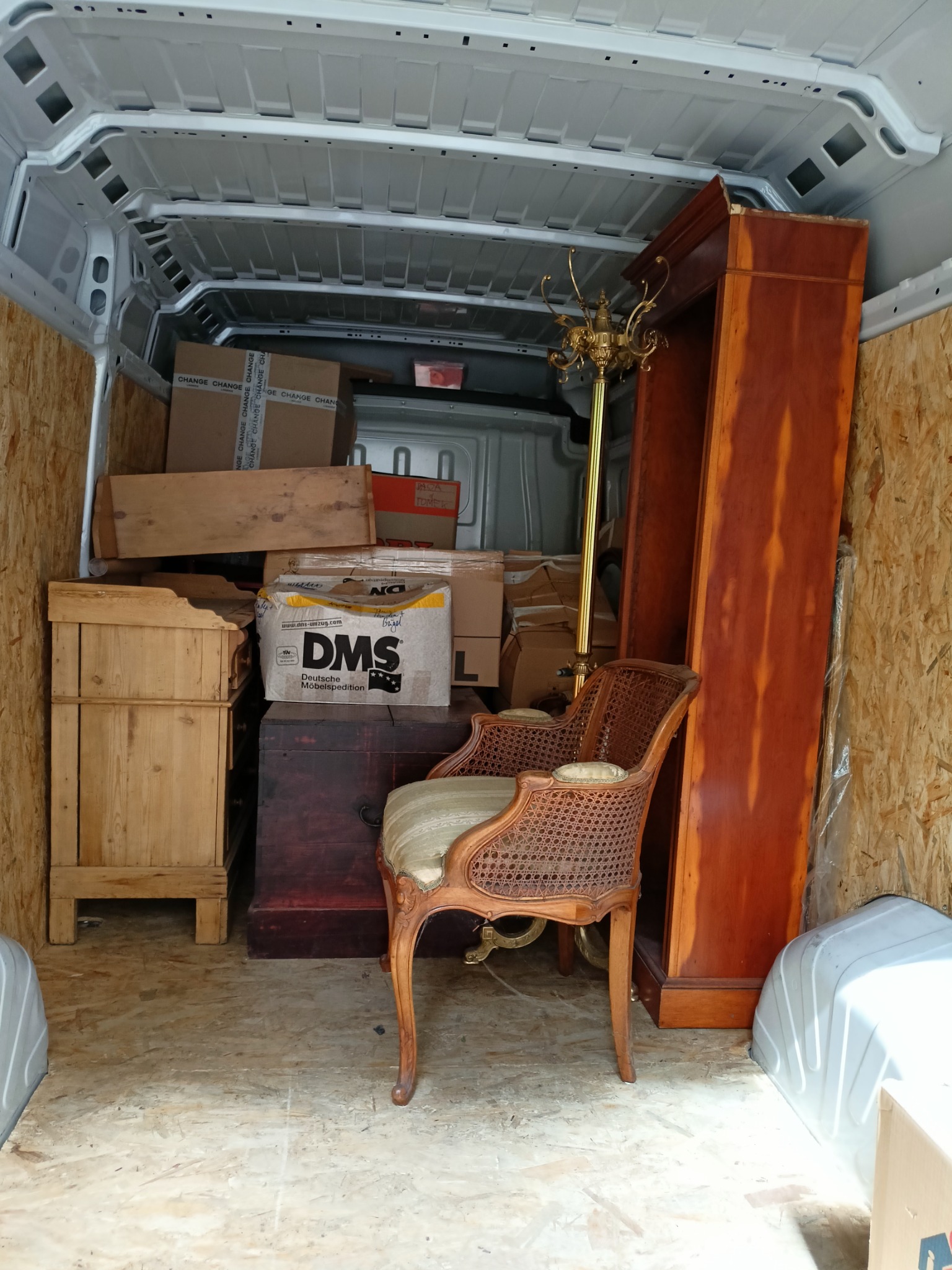 Wnętrze furgonetki załadowane meblami antycznymi, w tym krzesło z wiklinowym oparciem, drewniana komoda, wysoki regał i złoty wieszak na ubrania, oraz kartony z napisem 'DMS Deutsche Möbelspedition'.