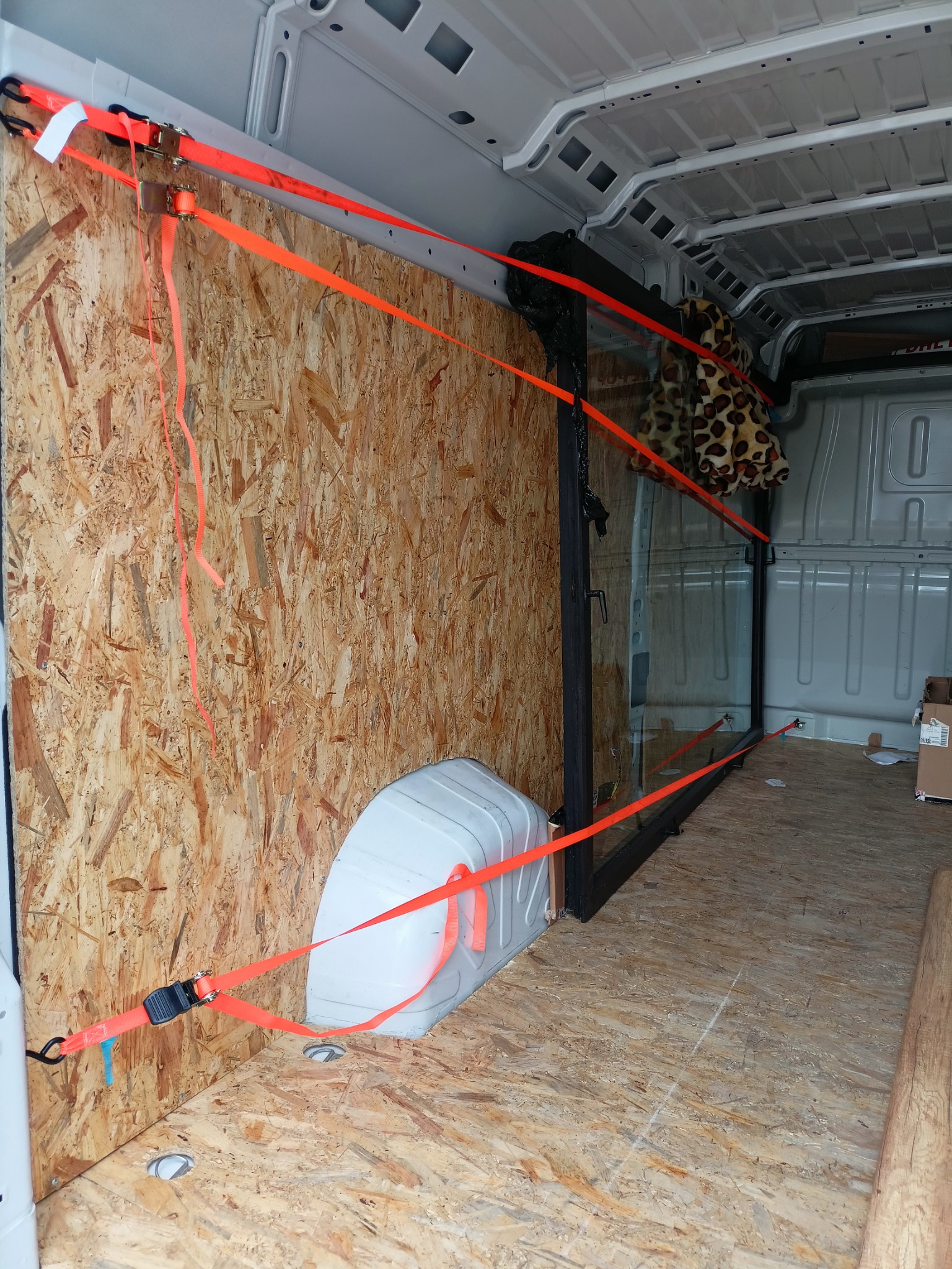 Wnętrze busa zabezpieczone sklejką i pasami transportowymi, przewożona szyba w metalowej ramie, widoczna pluszowa maskotka.