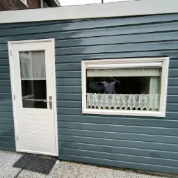 Świeżo pomalowany, drewniany budynek gospodarczy z białymi drzwiami i oknem z firanką. Widoczne ślady pędzla, podkreślające fakturę drewna i głębię koloru.