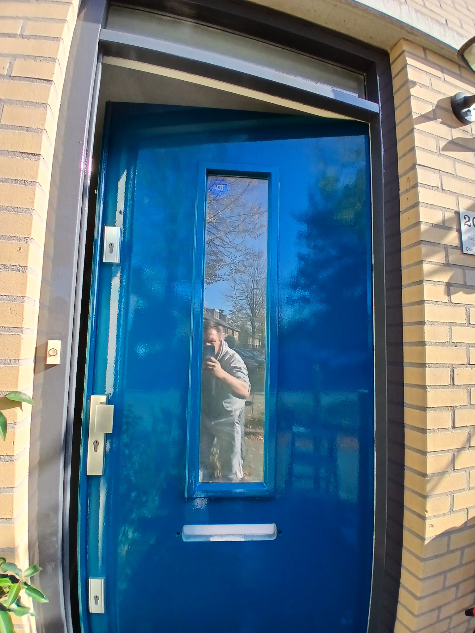 Błękitne, świeżo pomalowane drzwi wejściowe z szybką, z widocznym odbiciem osoby robiącej zdjęcie, na tle elewacji z jasnej cegły i szarej ramy.
