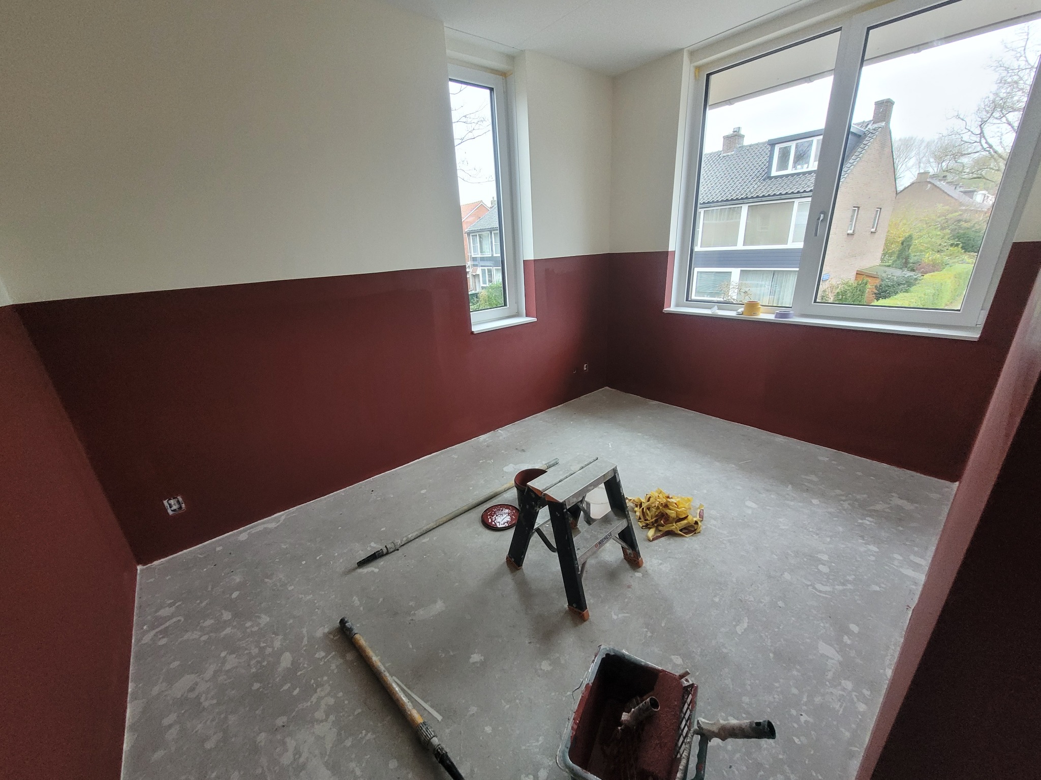 Pokój po częściowym malowaniu ścian na bordowo, widoczne narzędzia malarskie, drabina i okna z widokiem na zabudowę mieszkalną.