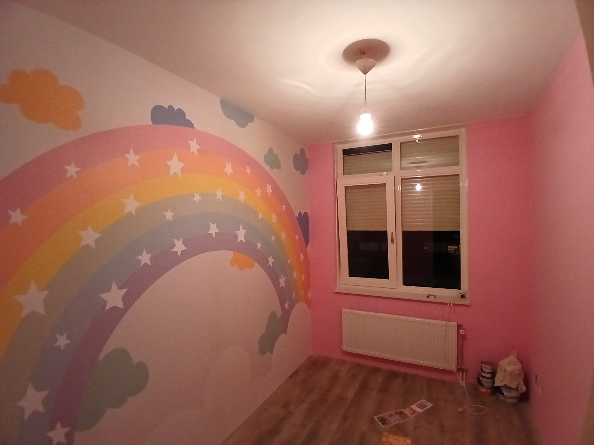 Pokój dziecięcy po malowaniu: tęcza z gwiazdami na ścianie, różowe ściany, białe okno i grzejnik. Widoczne akcesoria malarskie na podłodze.