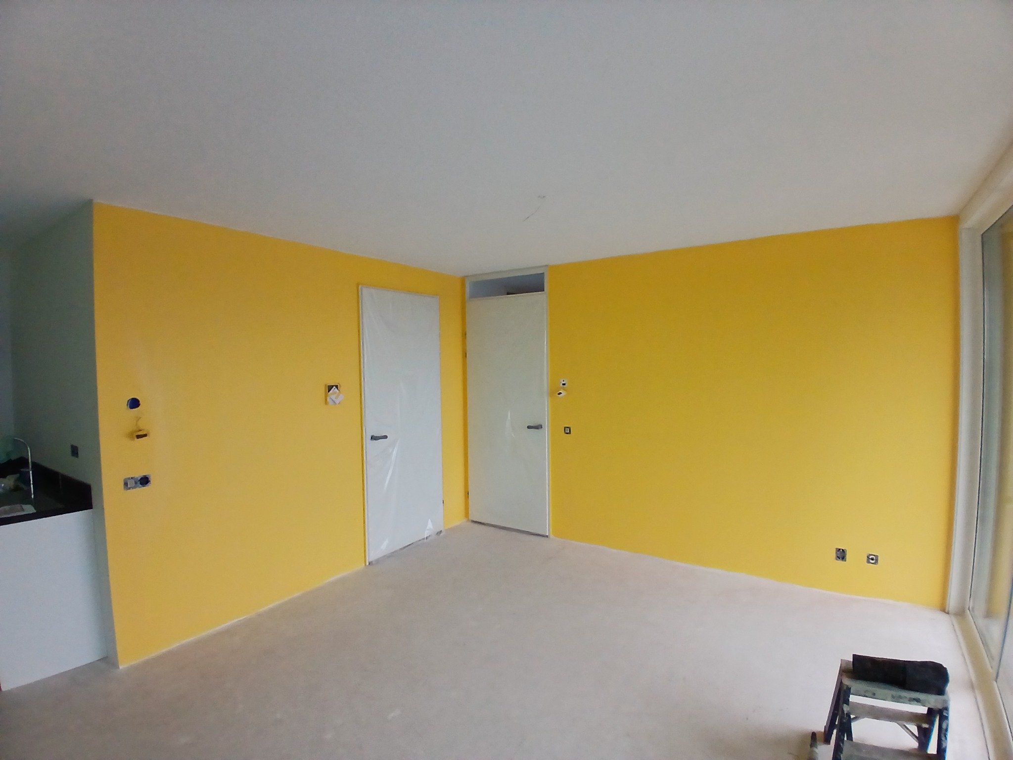 Żółte ściany w pokoju po malowaniu, zabezpieczone drzwi folią, drabina malarska w rogu. Widoczne gniazdka elektryczne. Jasne wnętrze z dużym oknem.
