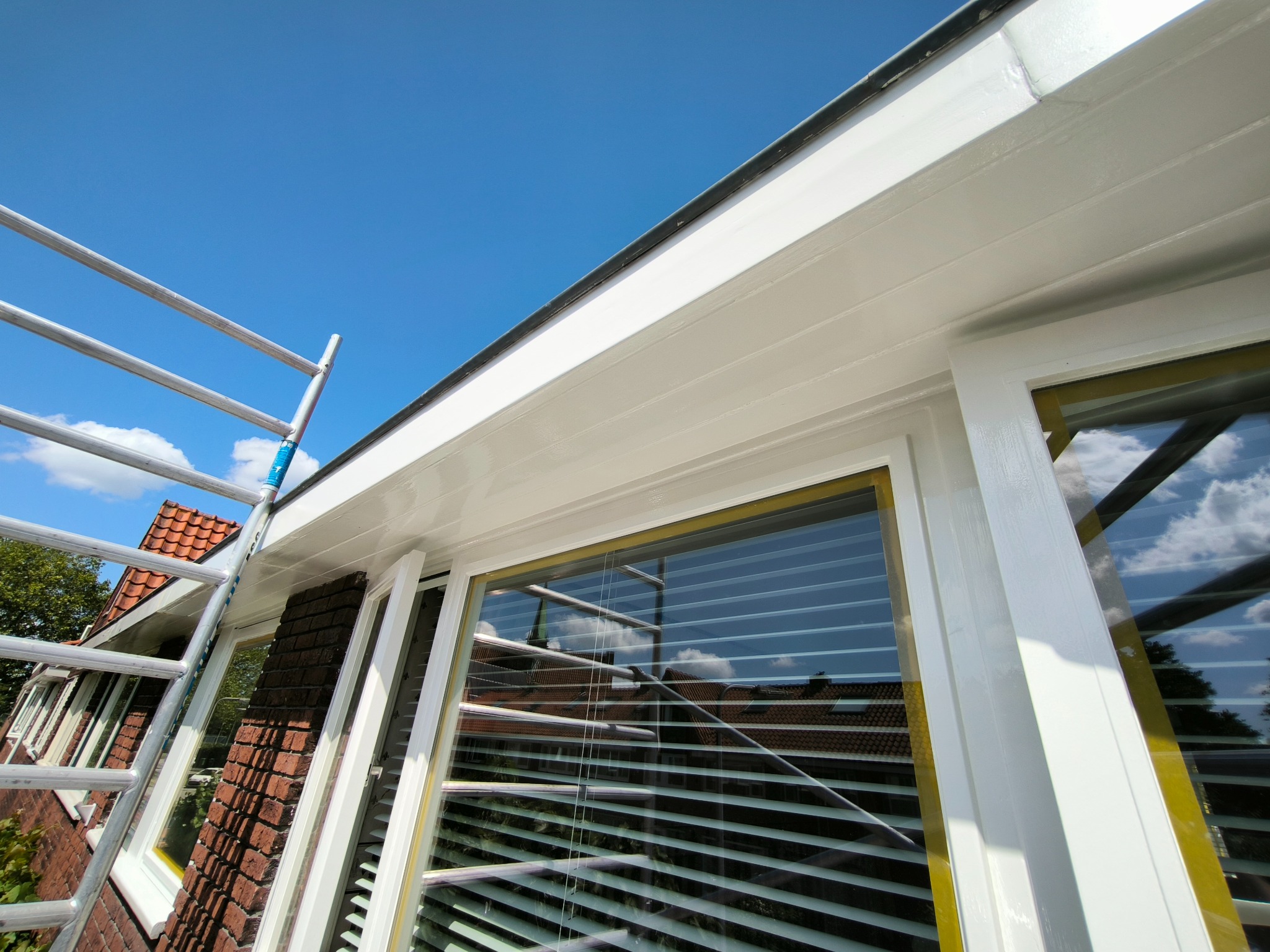 Białe, świeżo pomalowane okno z ramą i fragment elewacji domu z cegły, widoczne z dołu, z fragmentem drabiny malarskiej. Błękitne niebo w tle.