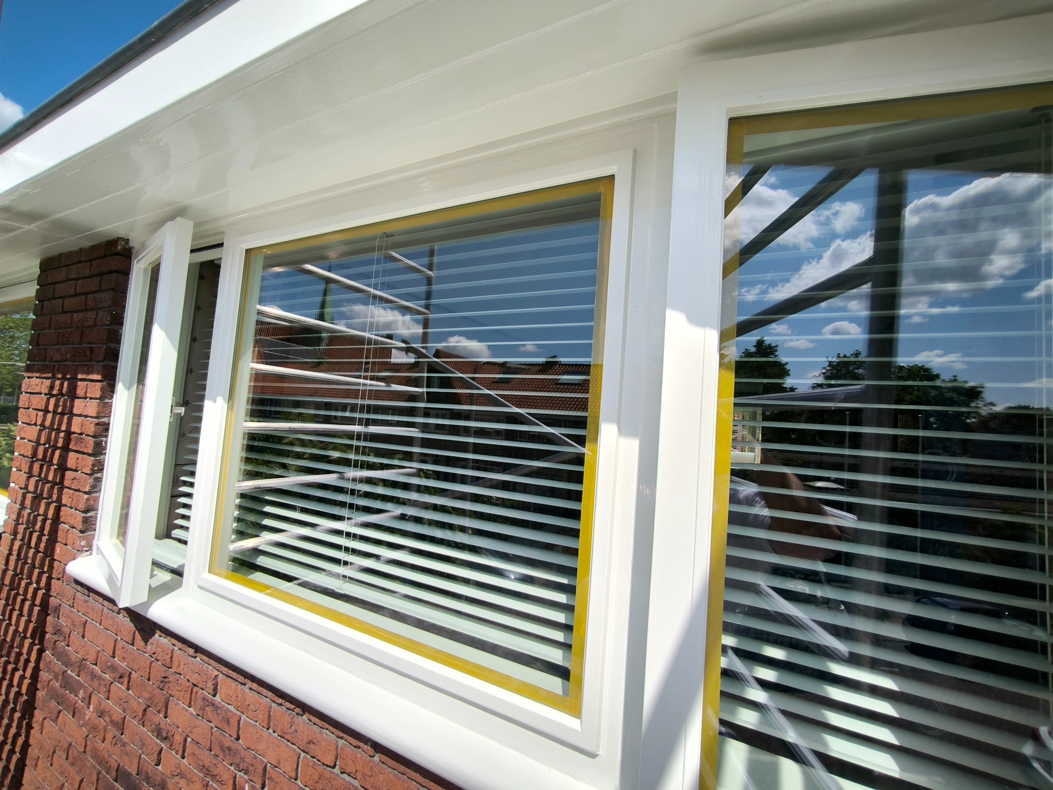 Białe, świeżo pomalowane okno z taśmą malarską w Delft, widoczne na tle ceglanej ściany i odbijającego się nieba w szybie. Otwarta część okna.