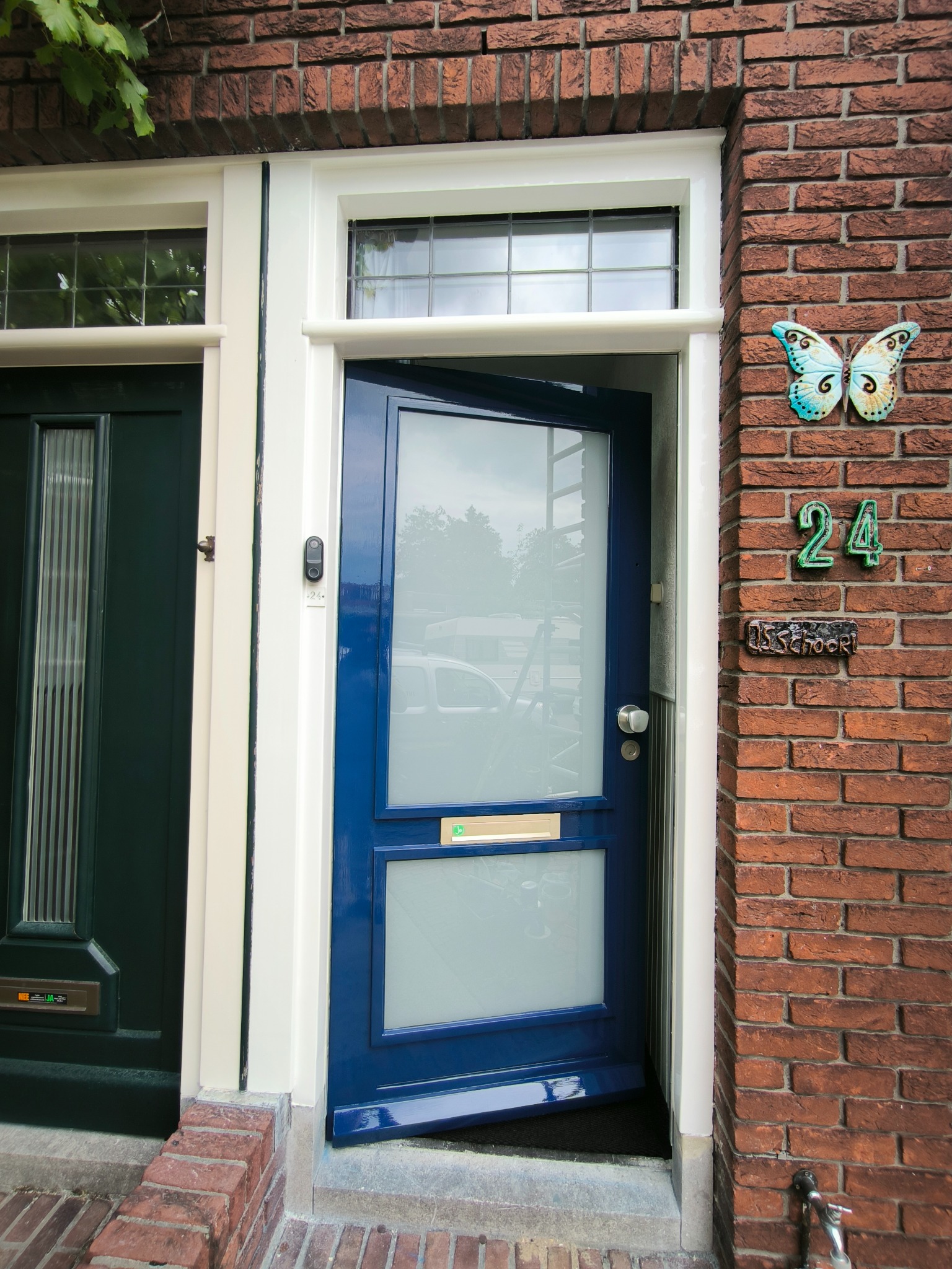 Niebieskie, częściowo otwarte drzwi z szybami w białej futrynie, na tle ceglanej ściany z numerem 24 i motylem. Obok zielone drzwi.