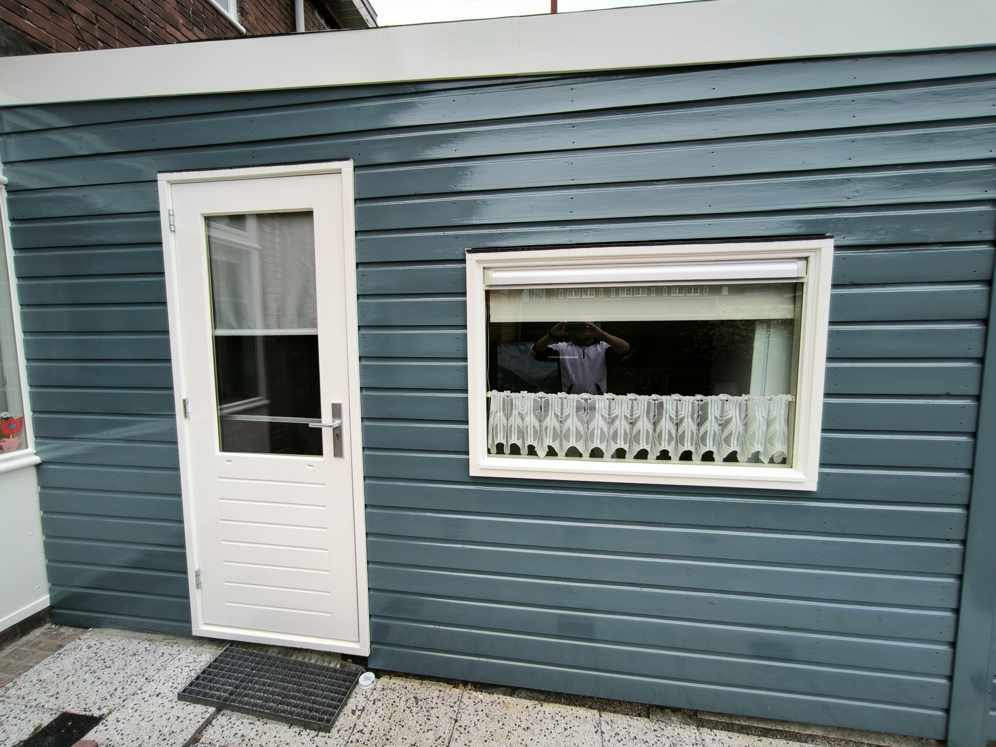 Świeżo pomalowany, drewniany budynek gospodarczy z białymi drzwiami i oknem z firanką. Widoczne ślady pędzla, podkreślające fakturę drewna i głębię koloru.