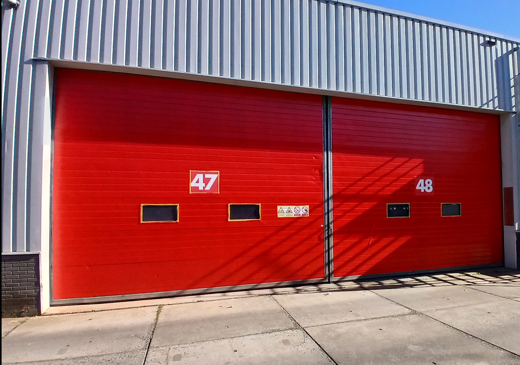 Czerwona, segmentowa brama garażowa z numerami 47 i 48, z małymi okienkami, na tle szarej, blaszanej elewacji budynku przemysłowego w słoneczny dzień.