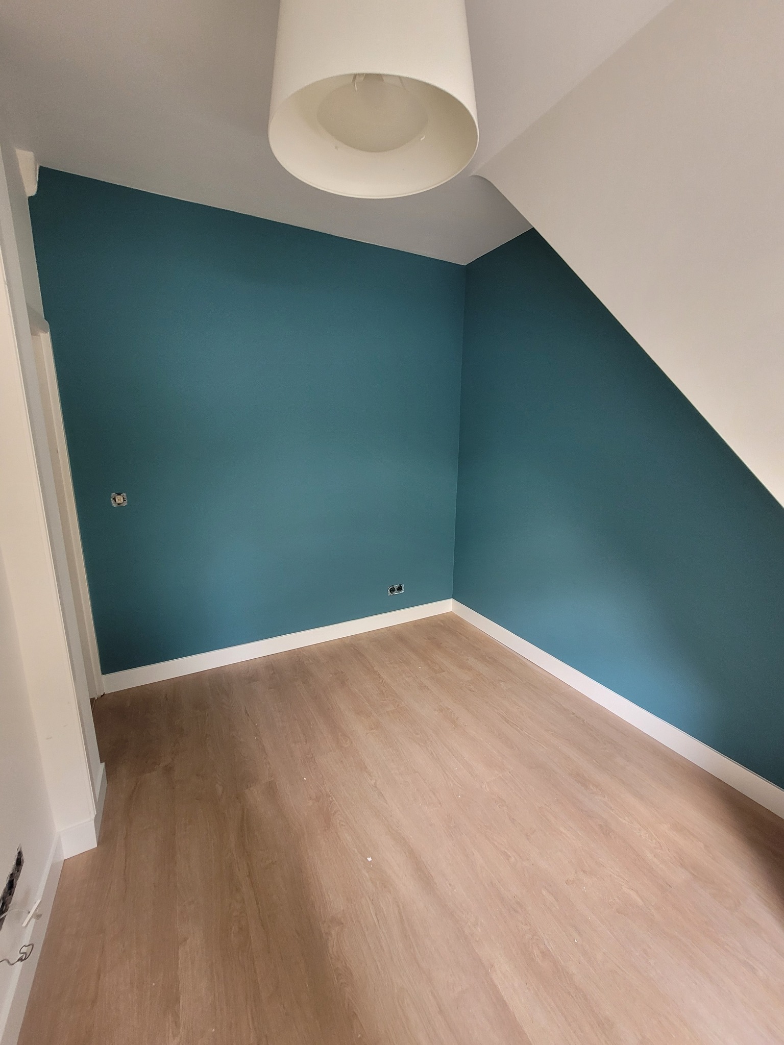 Pokój po remoncie: ściany pomalowane na turkusowo, białe listwy przypodłogowe, jasna podłoga laminowana, biały sufit ze skośnym fragmentem i biała lampa sufitowa.