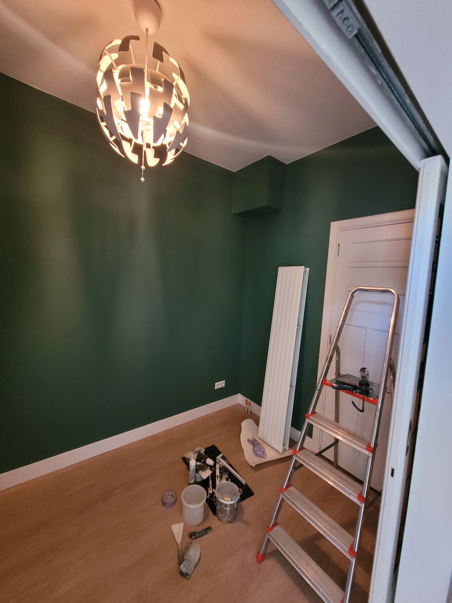 Pokój po malowaniu na ciemnozielony kolor, widoczna drabina, grzejnik oparty o ścianę, lampa sufitowa w stylu skandynawskim i akcesoria malarskie na podłodze.