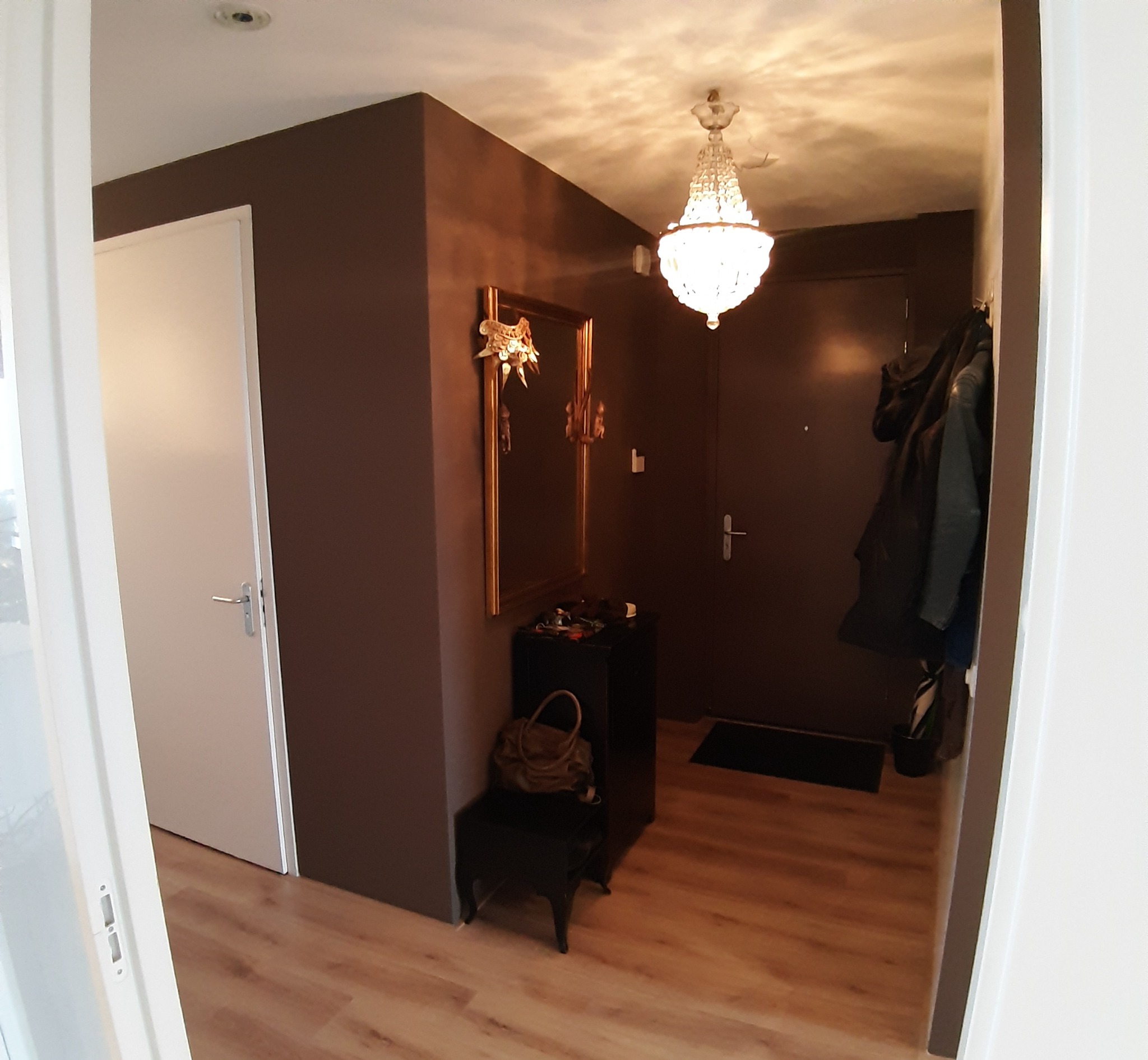 Przedpokój z brązowymi ścianami, lustrem w złotej ramie, kryształowym żyrandolem i drewnianą podłogą.