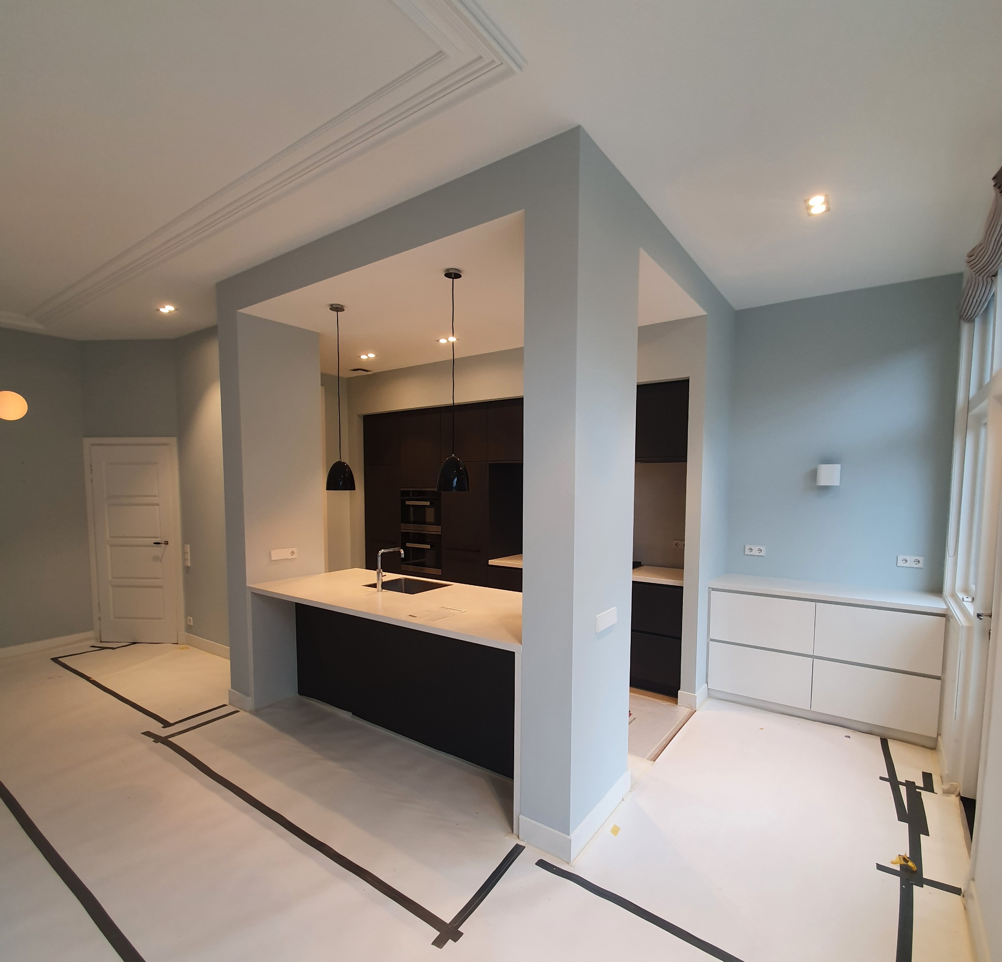 Wnętrze po remoncie: Jasnoszare ściany, nowoczesna czarna zabudowa kuchenna z wyspą i białym blatem, zabezpieczona podłoga z taśmą malarską, minimalistyczne oświetlenie.