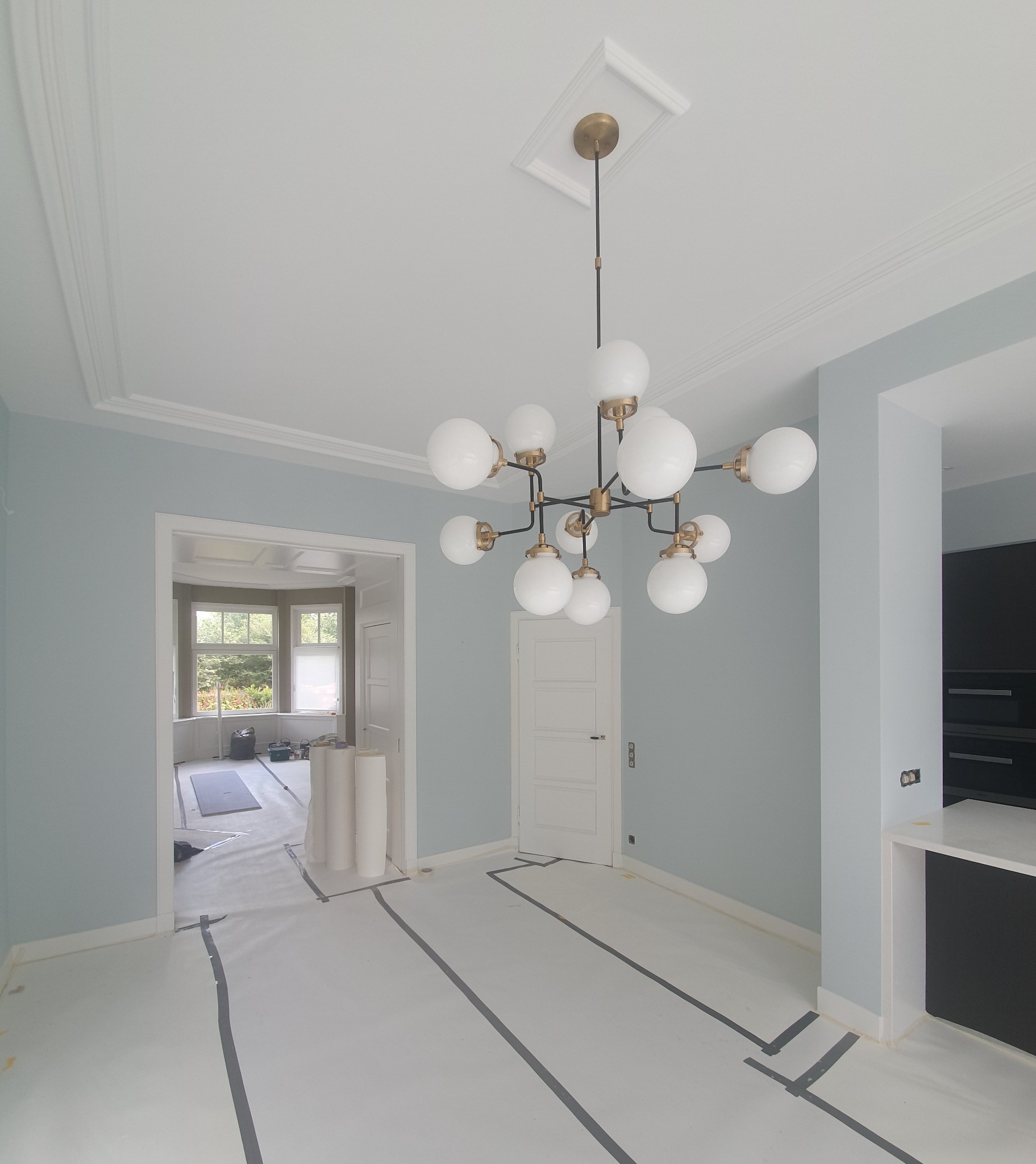 Przygotowane do malowania wnętrze w Delft, z zabezpieczoną folią podłogą i ścianami pomalowanymi na jasny odcień niebieskiego, widoczny nowoczesny żyrandol.