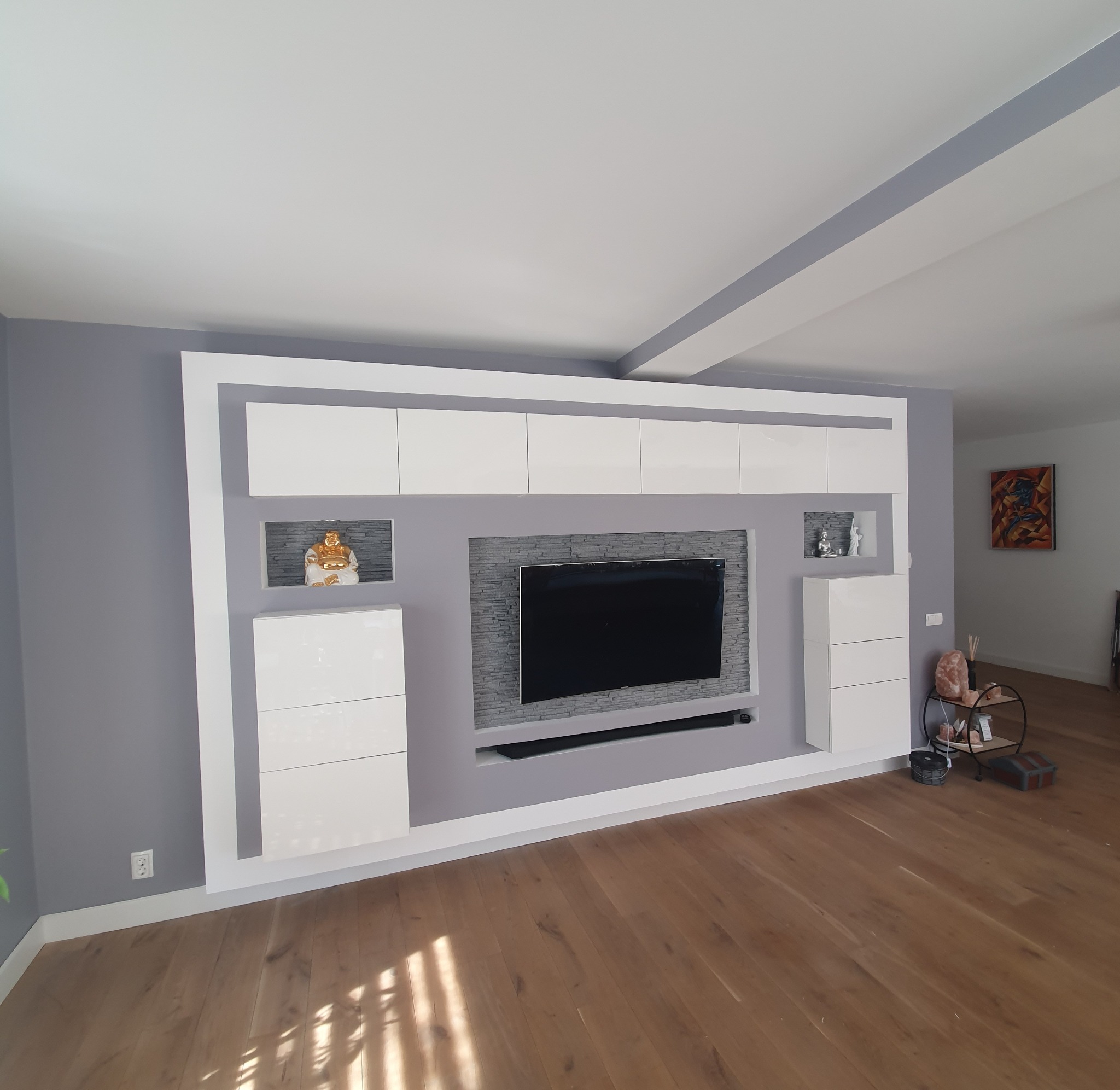 Szara ściana w salonie z białą zabudową meblową, w której wkomponowano telewizor i dekoracyjne nisze z figurkami Buddy, drewniana podłoga.