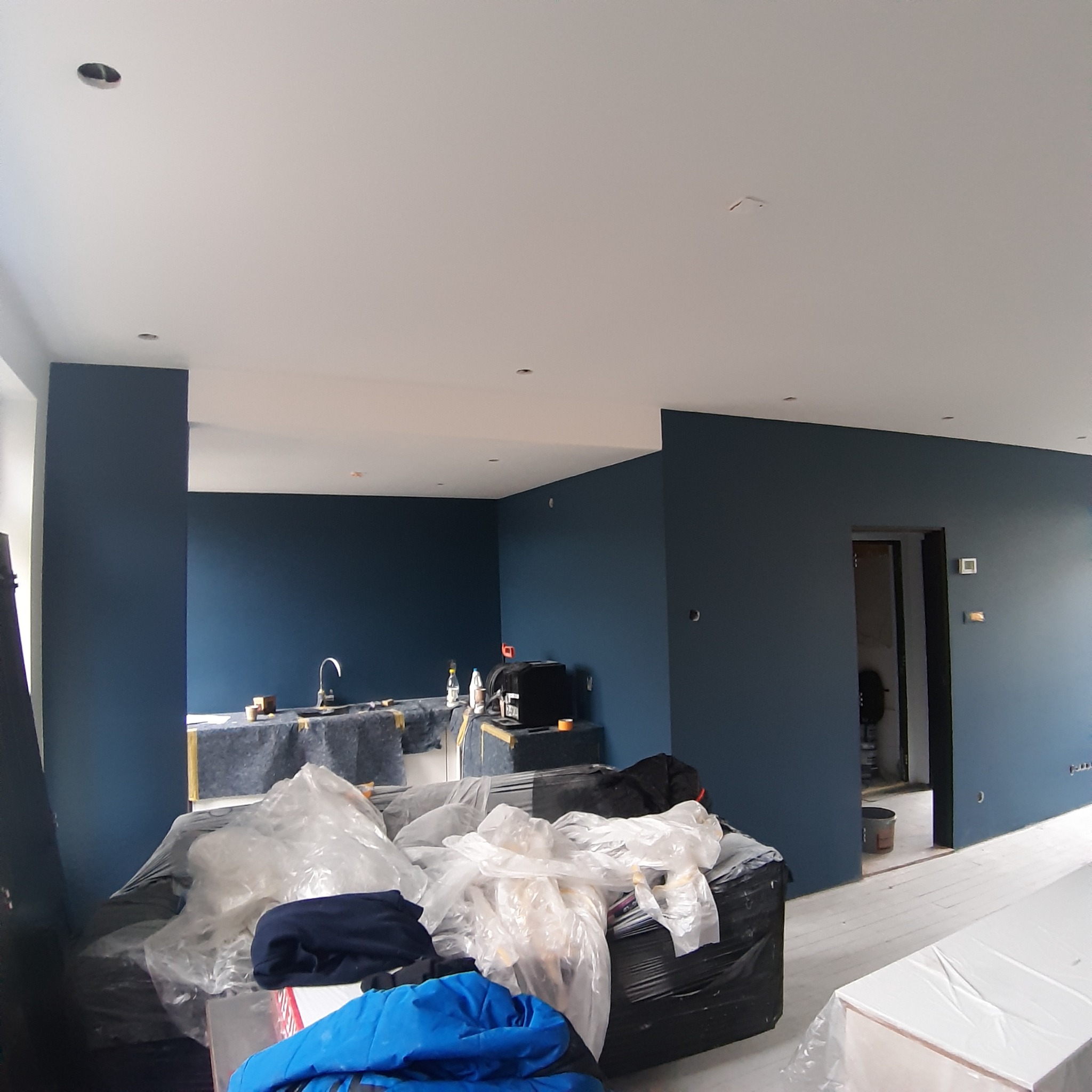 Pokój w trakcie remontu, ściany pomalowane na ciemny niebieski kolor, meble i podłoga zabezpieczone folią malarską, widoczny zlew i akcesoria w tle.