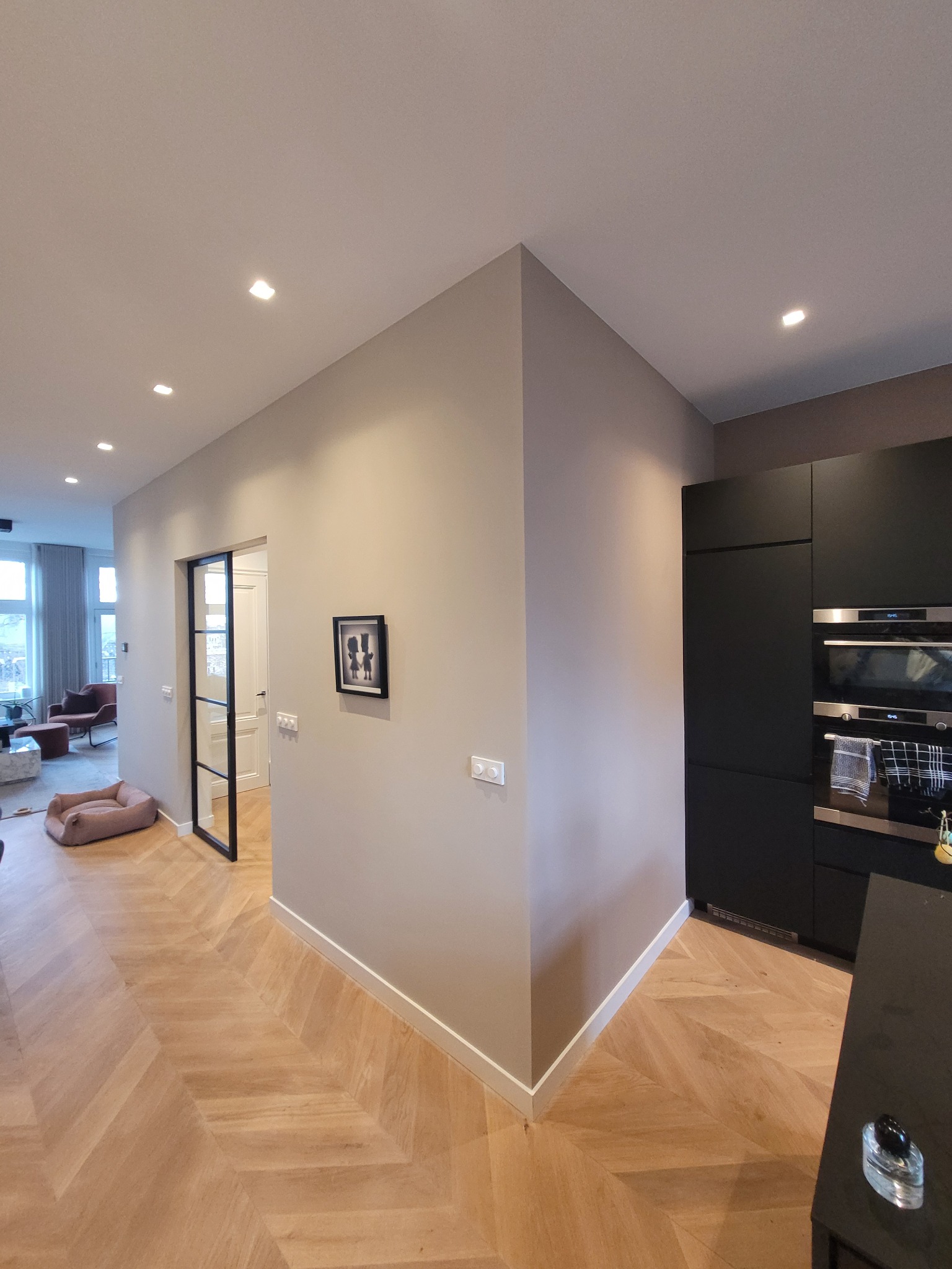 Jasnoszara ściana po remoncie, z zamontowanymi włącznikami i czarną ramką ze zdjęciem, widok na salon i kuchnię z parkietem ułożonym w jodełkę.