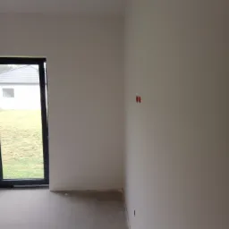 Świeżo pomalowane na biało wnętrze pokoju z widocznymi gniazdkami elektrycznymi bez osłon, szarym wykończeniem podłogi i oknem z widokiem na dom i trawnik.