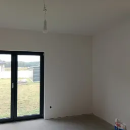 Pokój po świeżo wykonanych pracach wykończeniowych: gładkie, pomalowane na biało ściany, szara wylewka podłogowa, widoczne okno balkonowe z czarną ramą i grzejnik. Na suficie wisi żarówka na kablu.