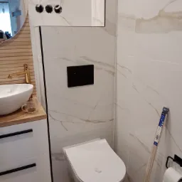 Nowoczesna łazienka w trakcie wykańczania: białe płytki imitujące marmur, podwieszana toaleta z czarnym przyciskiem, umywalka nablatowa z złotą armaturą i drewnianym blatem, widoczne przewody...