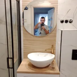 Nowoczesna łazienka z okrągłym lustrem, w którym odbija się mężczyzna robiący zdjęcie telefonem, widoczna biała umywalka nablatowa, złota bateria, dekoracyjne płytki imitujące drewno na ścianie...