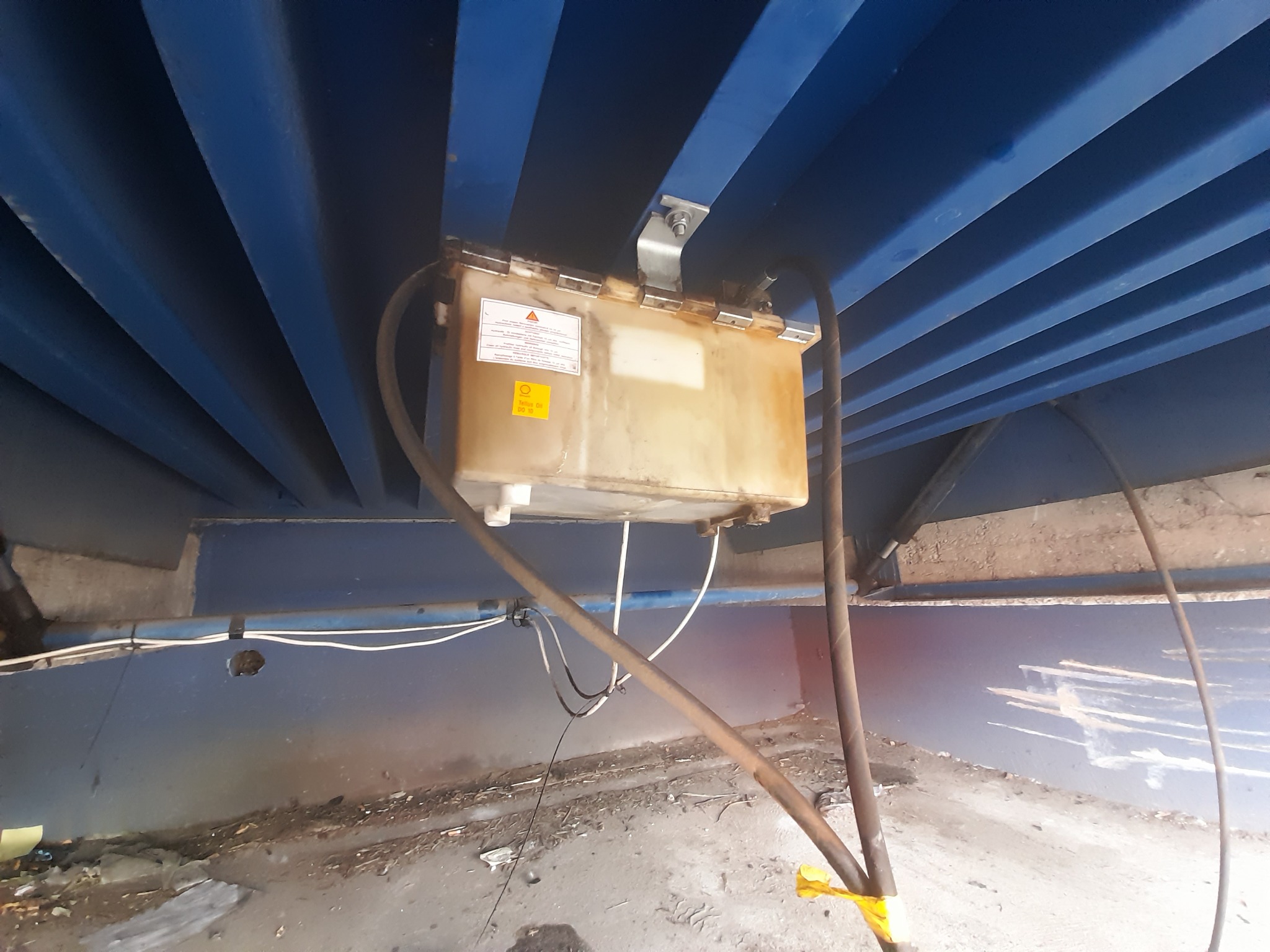 Zbiornik z olejem hydraulicznym zamontowany pod konstrukcją bramy przemysłowej, widoczne przewody i mocowanie.