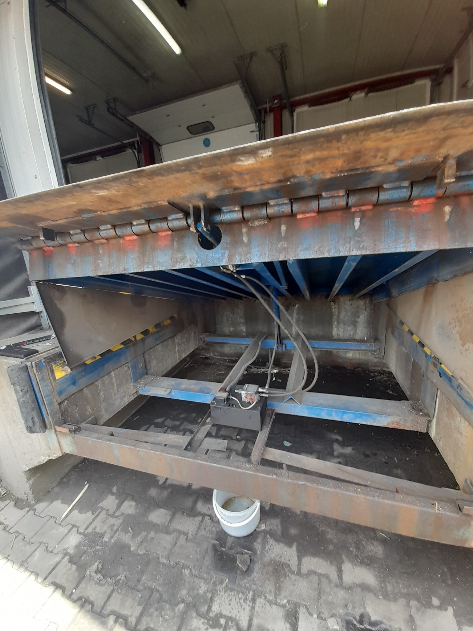 Otwarta rampa załadunkowa z widocznymi elementami hydraulicznymi i konstrukcją stalową, zarys wnętrza magazynu w tle.