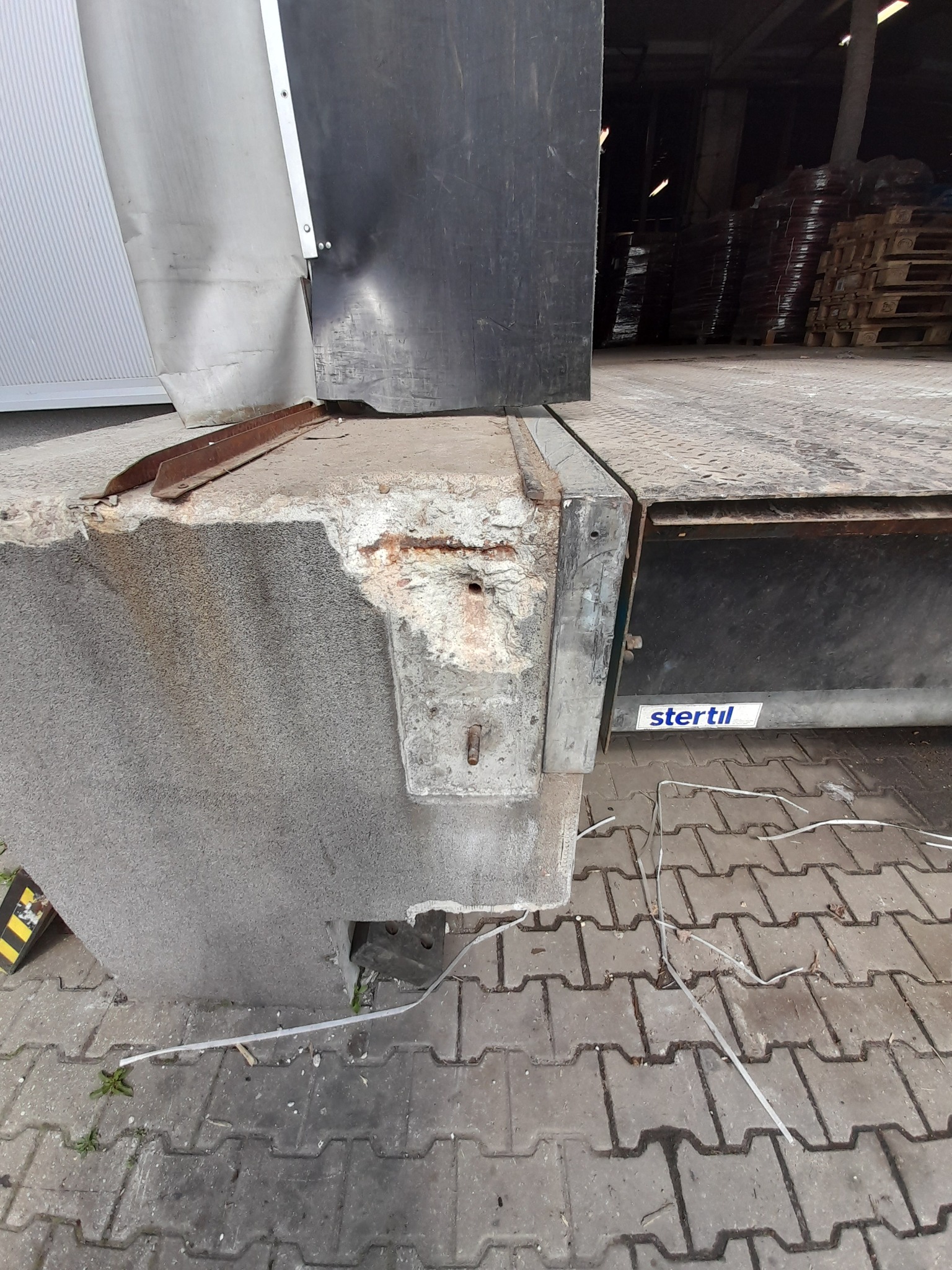 Uszkodzony betonowy najazd rampy załadunkowej z widocznymi śladami korozji i ubytkami, obok brama przemysłowa z logo Stertil.