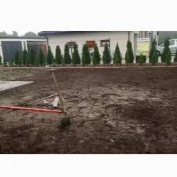 Przygotowany teren pod trawnik z narzędziami: łopata wbita w ziemię, grabie i poziomica leżące obok, w tle rząd tuj i budynki.